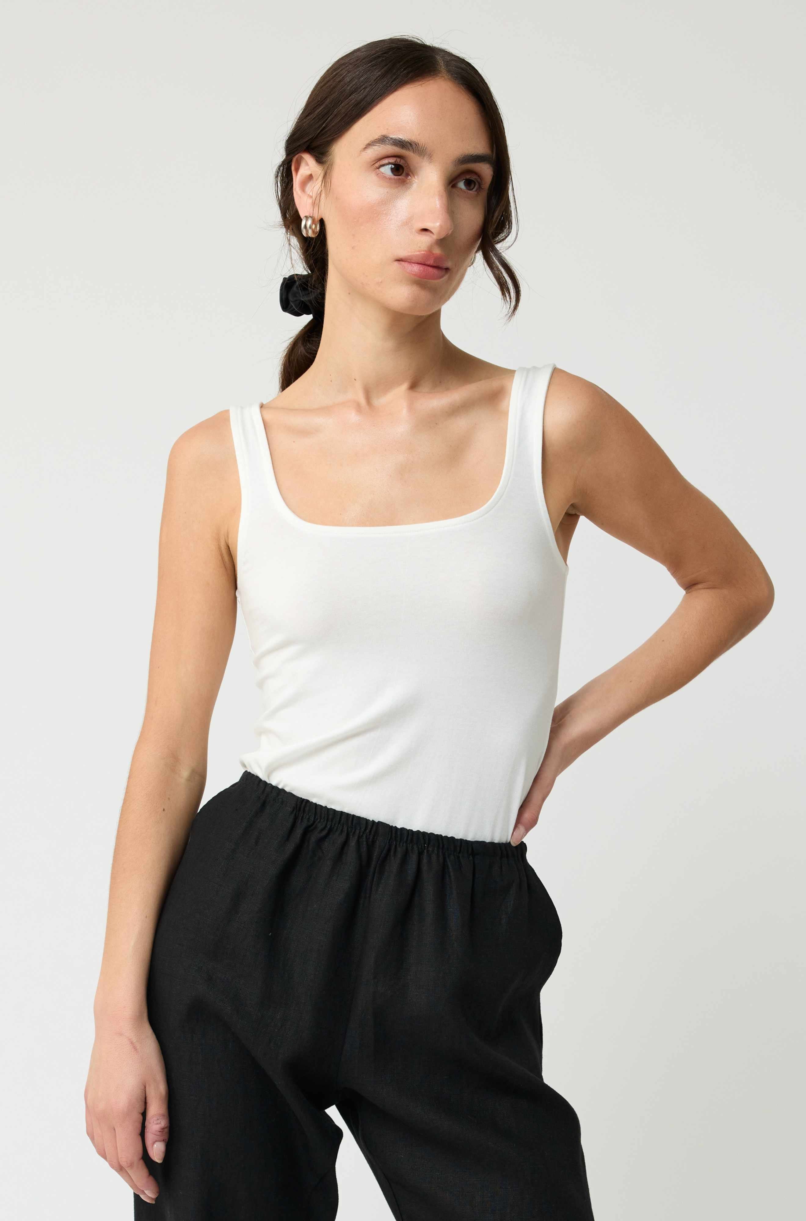 Woman wearing a white tank top and black pants against a plain background