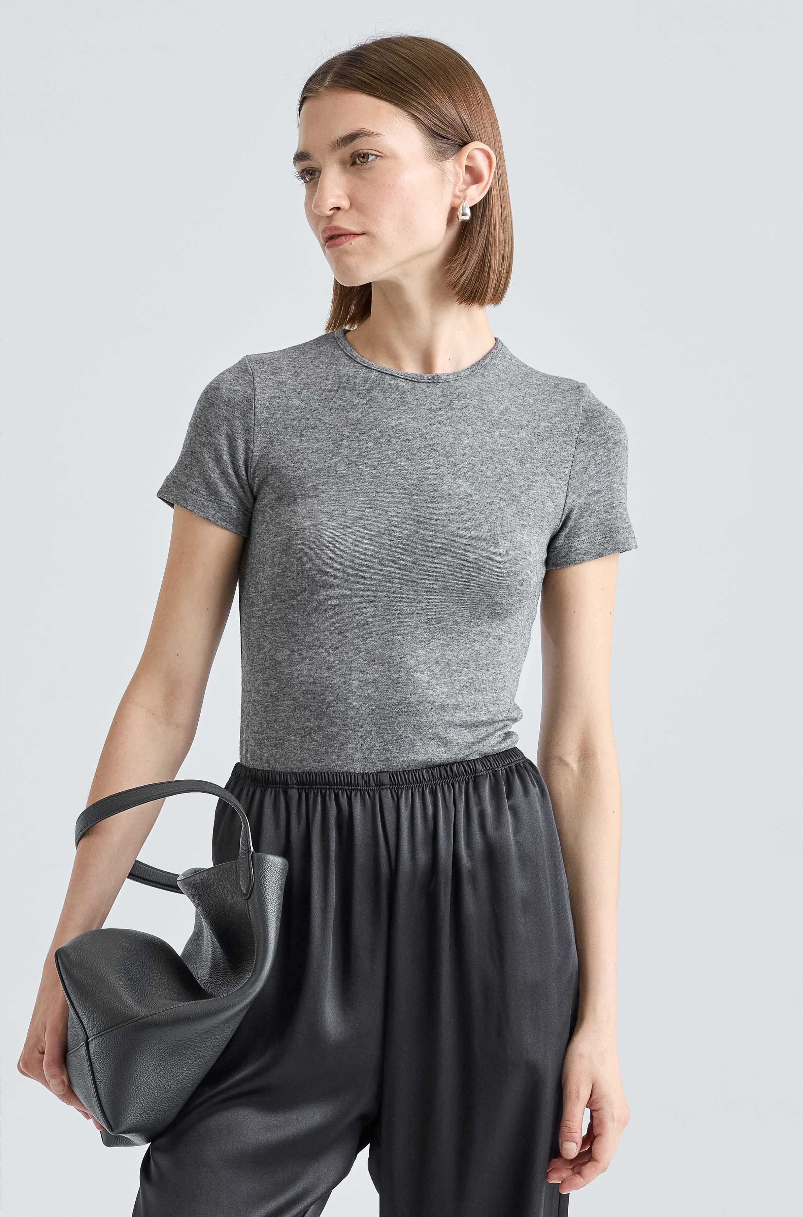 Woman wearing a gray t-shirt and black pants holding a black handbag against a light gray background