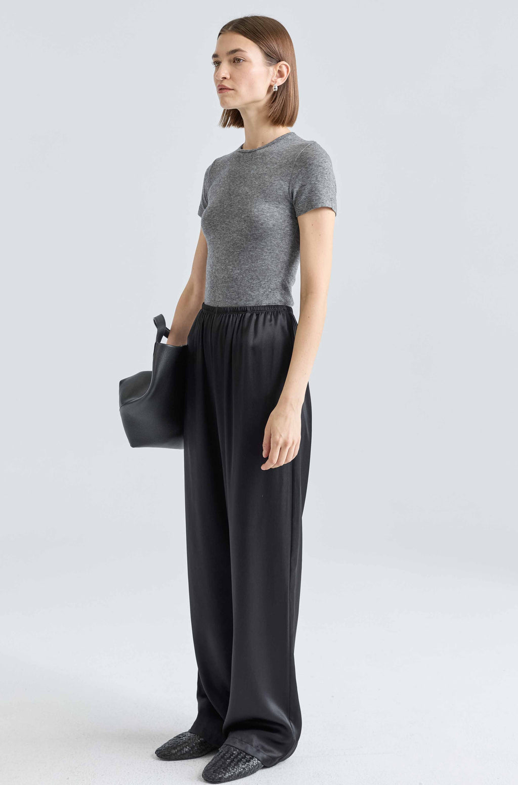 Woman wearing a gray sweater and black pants holding a black handbag on a light gray background
