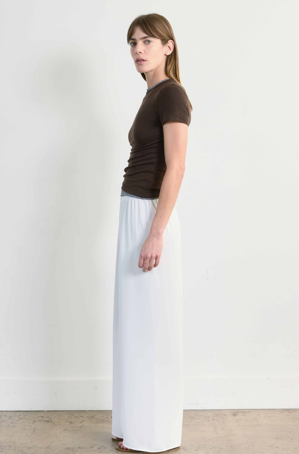 Woman wearing a brown t-shirt and white pants standing against a plain white wall.