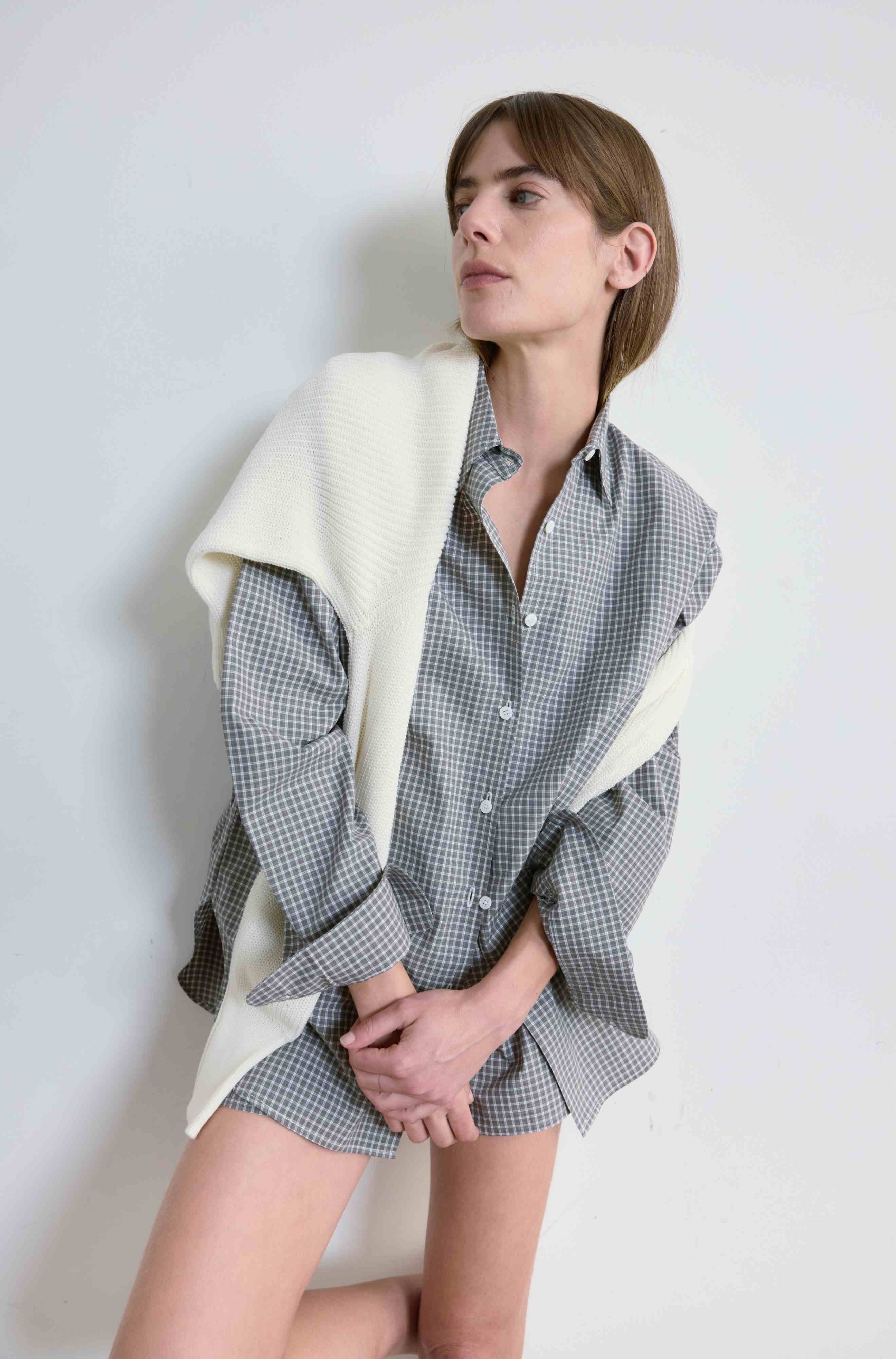 Woman wearing a checkered shirt and shorts with a white blanket draped over her shoulders against a plain background