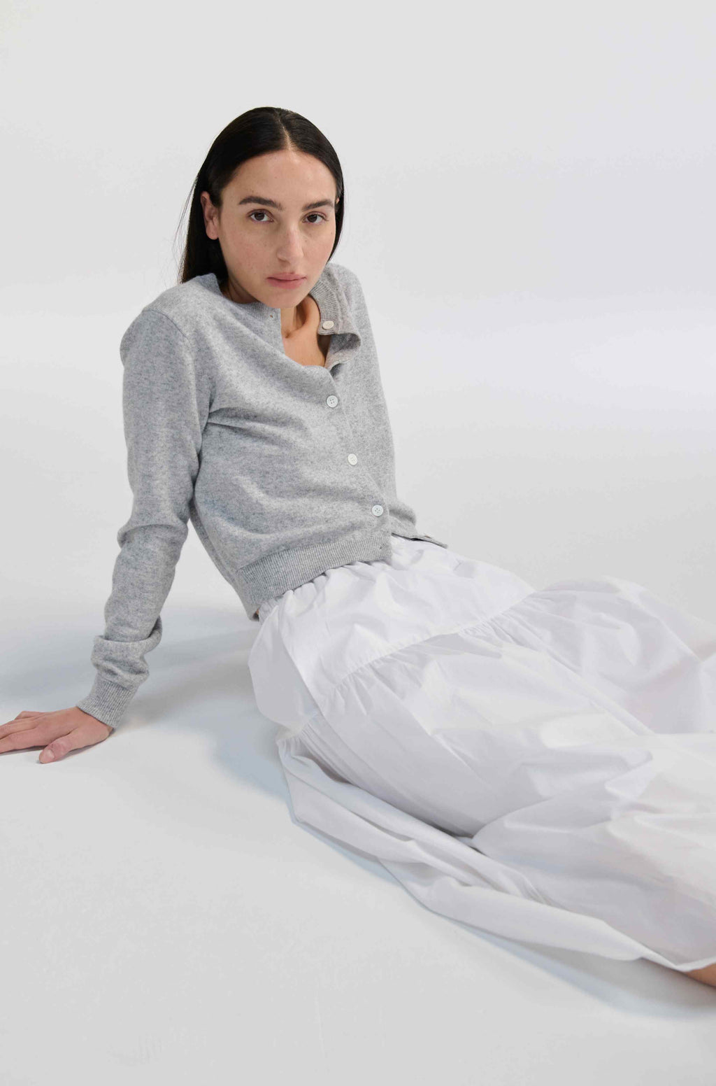 Woman wearing a gray cardigan and white skirt sitting on a white surface.
