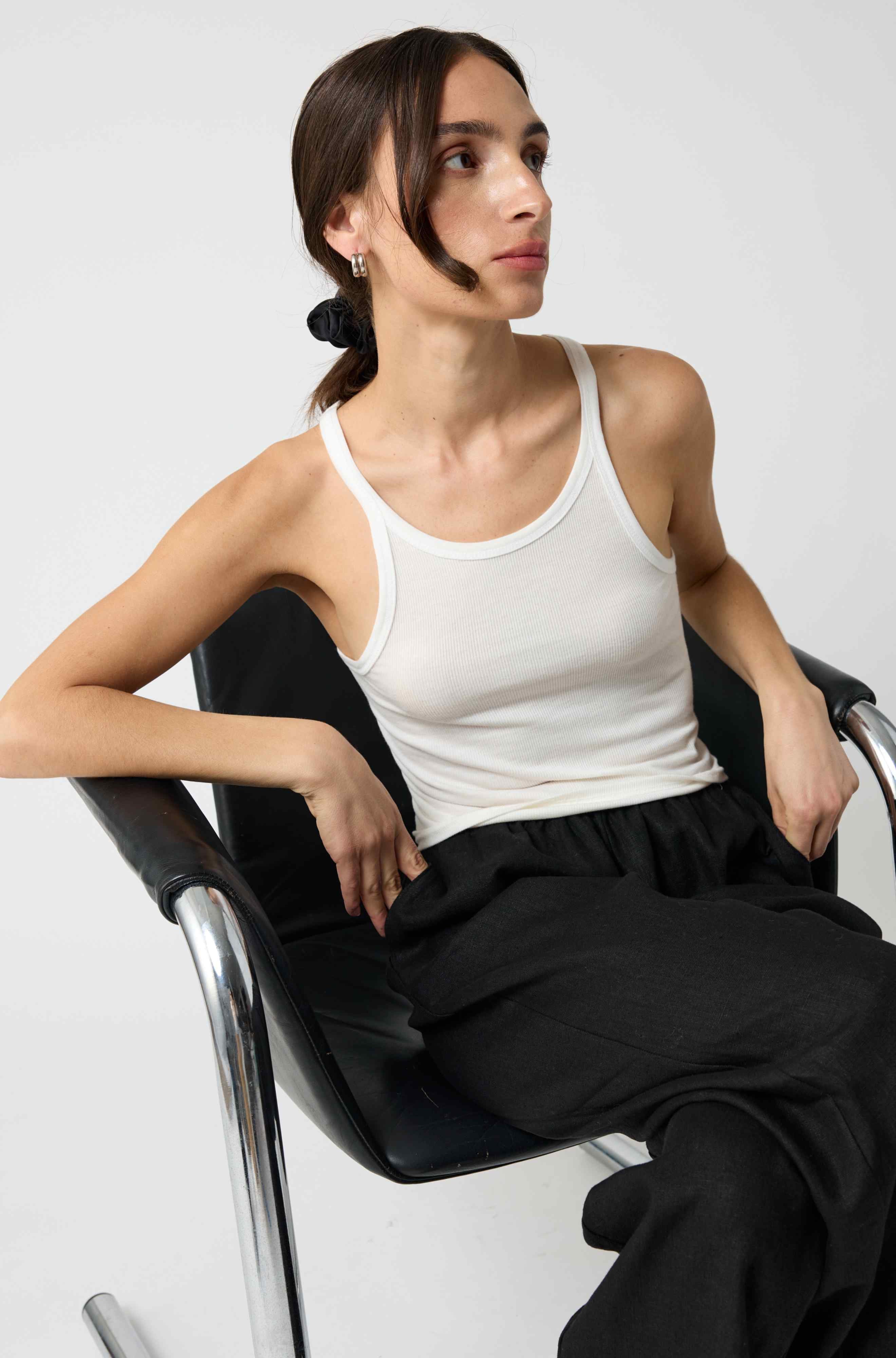 Person sitting in a chair wearing a white tank top and black pants on a plain background