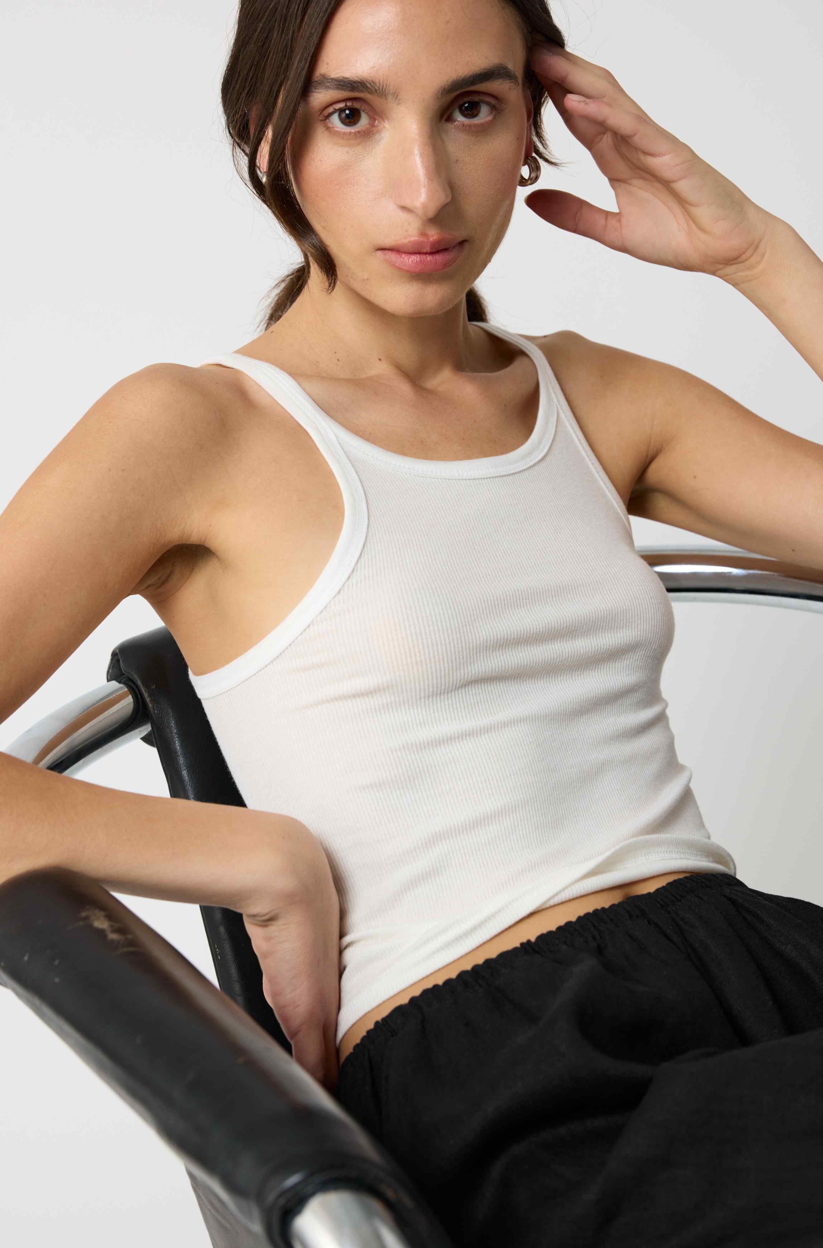 Woman wearing a white tank top and black pants sitting on a chair against a plain background