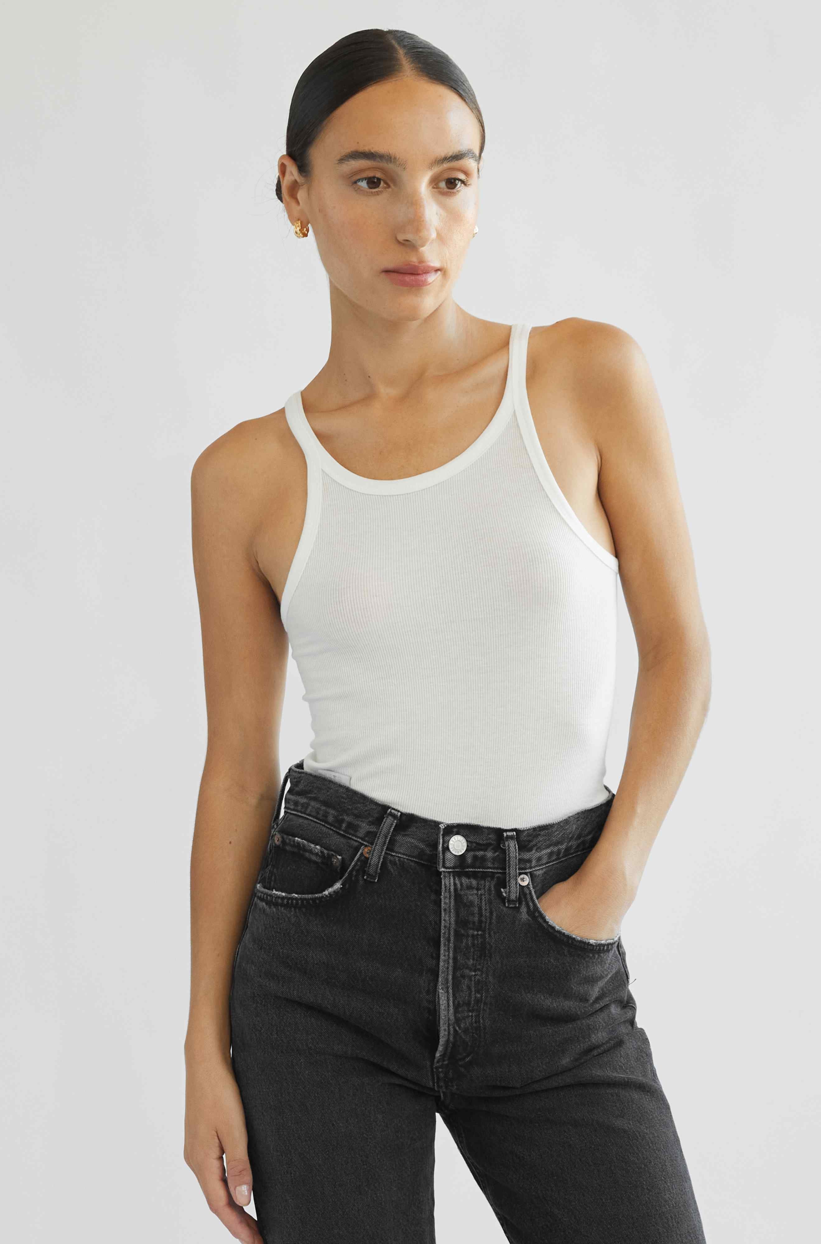 Woman wearing a white tank top and black jeans against a plain background