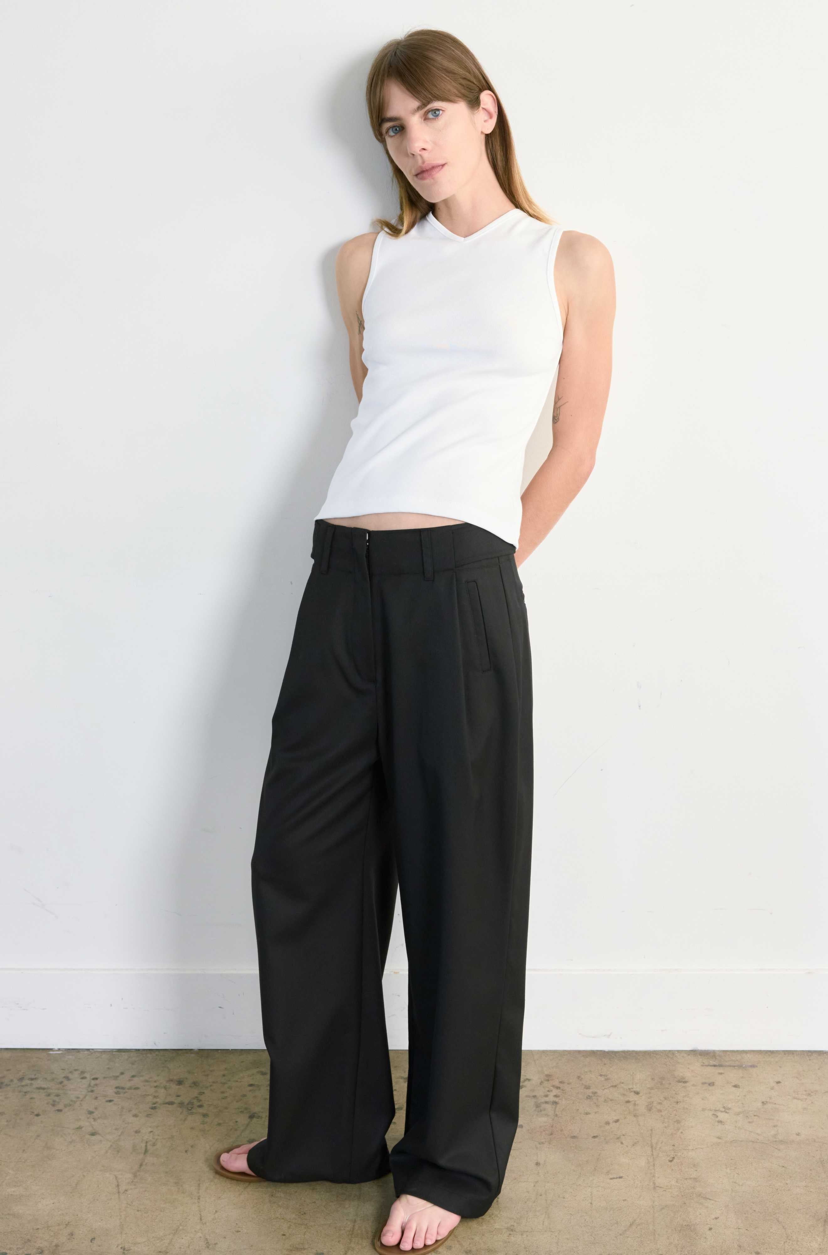 Woman wearing a white sleeveless top and black wide-leg pants against a white wall.
