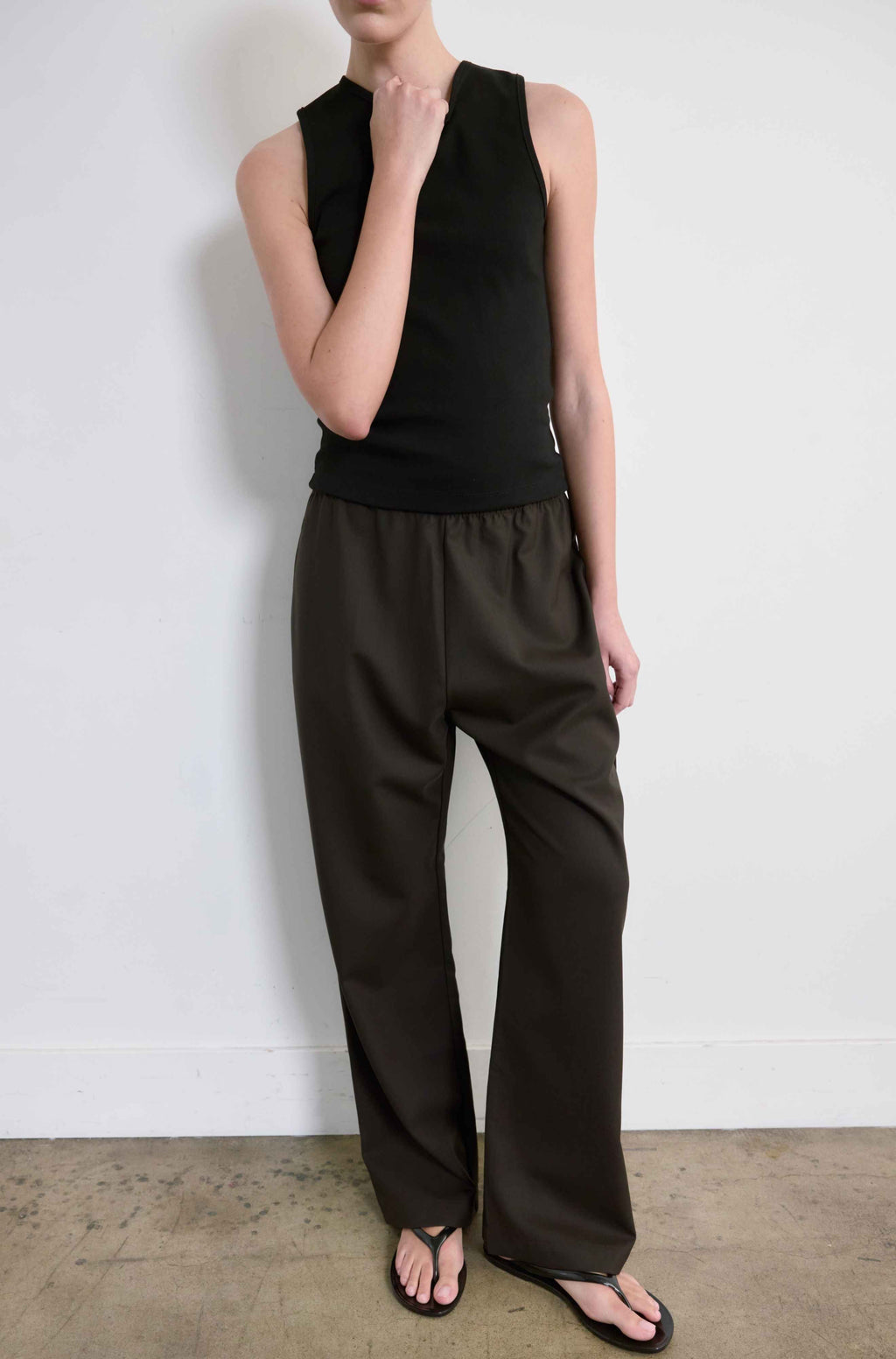 Person wearing a black sleeveless top and brown pants against a white wall.