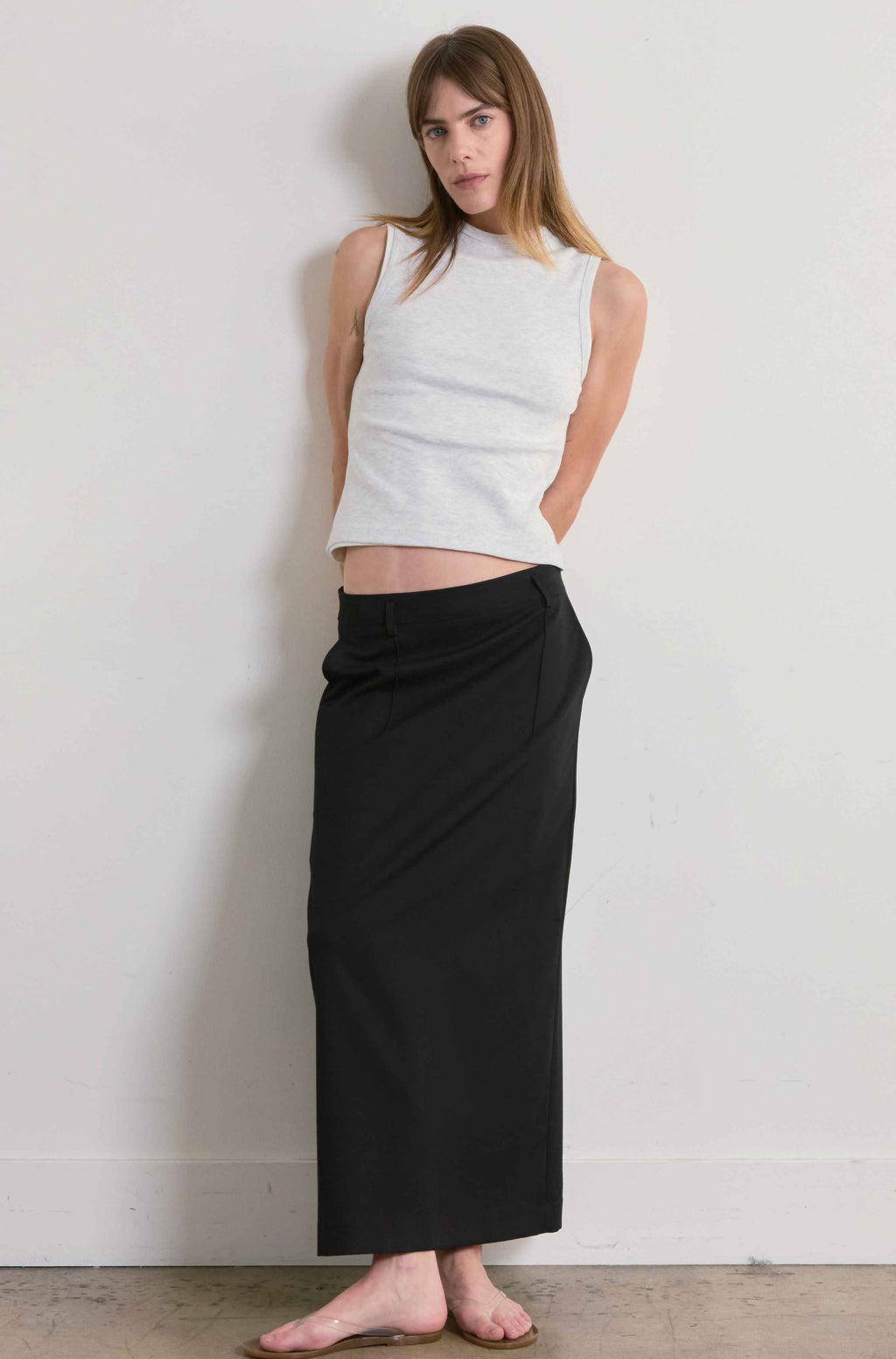 Woman wearing a grey sleeveless top and black skirt against a plain wall.
