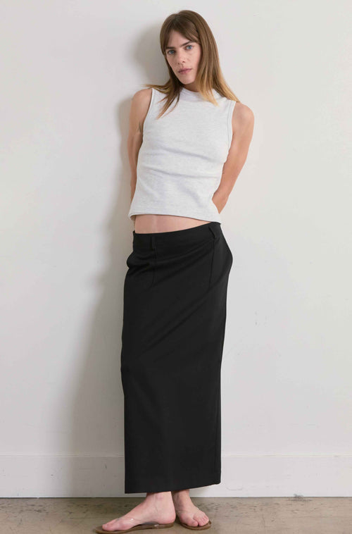 Woman wearing a grey sleeveless top and black skirt against a plain wall.
