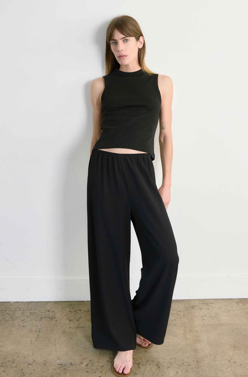 Woman wearing a black sleeveless top and wide-leg pants against a white wall.