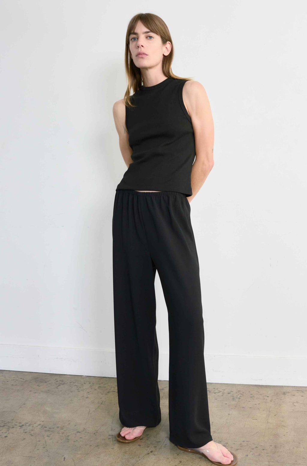 Woman wearing a black sleeveless top and wide-leg pants against a white wall.
