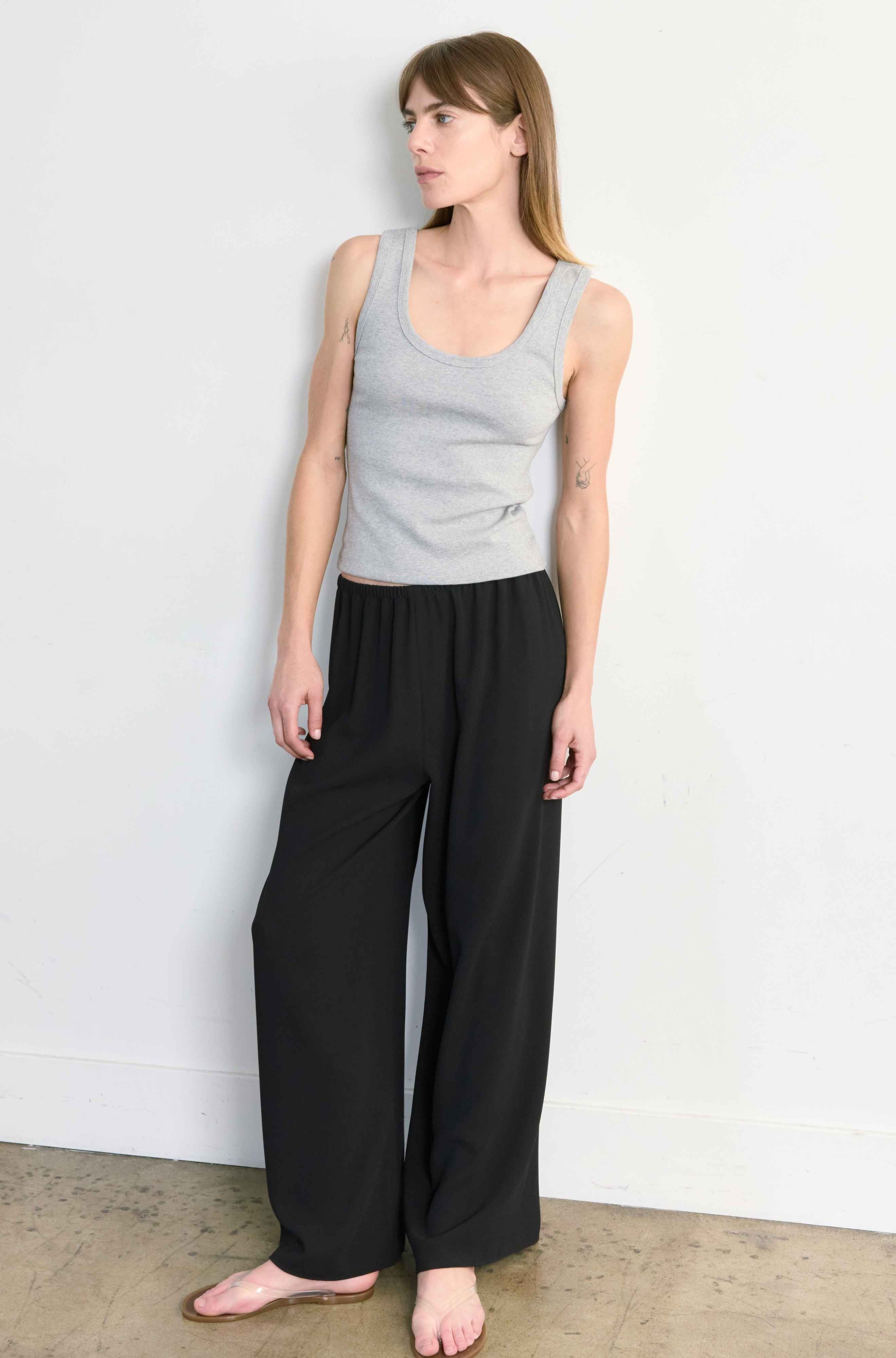 Woman wearing a gray tank top and black pants against a white wall.