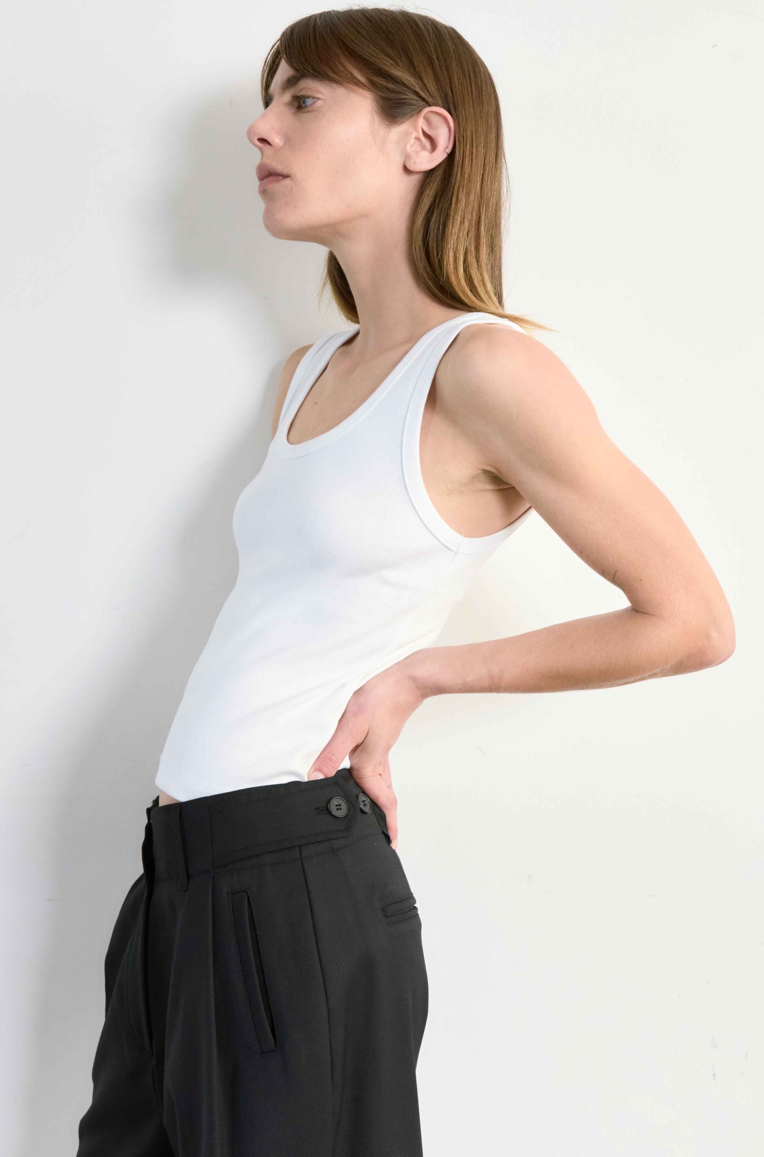 Woman wearing a white tank top and black pants against a plain background