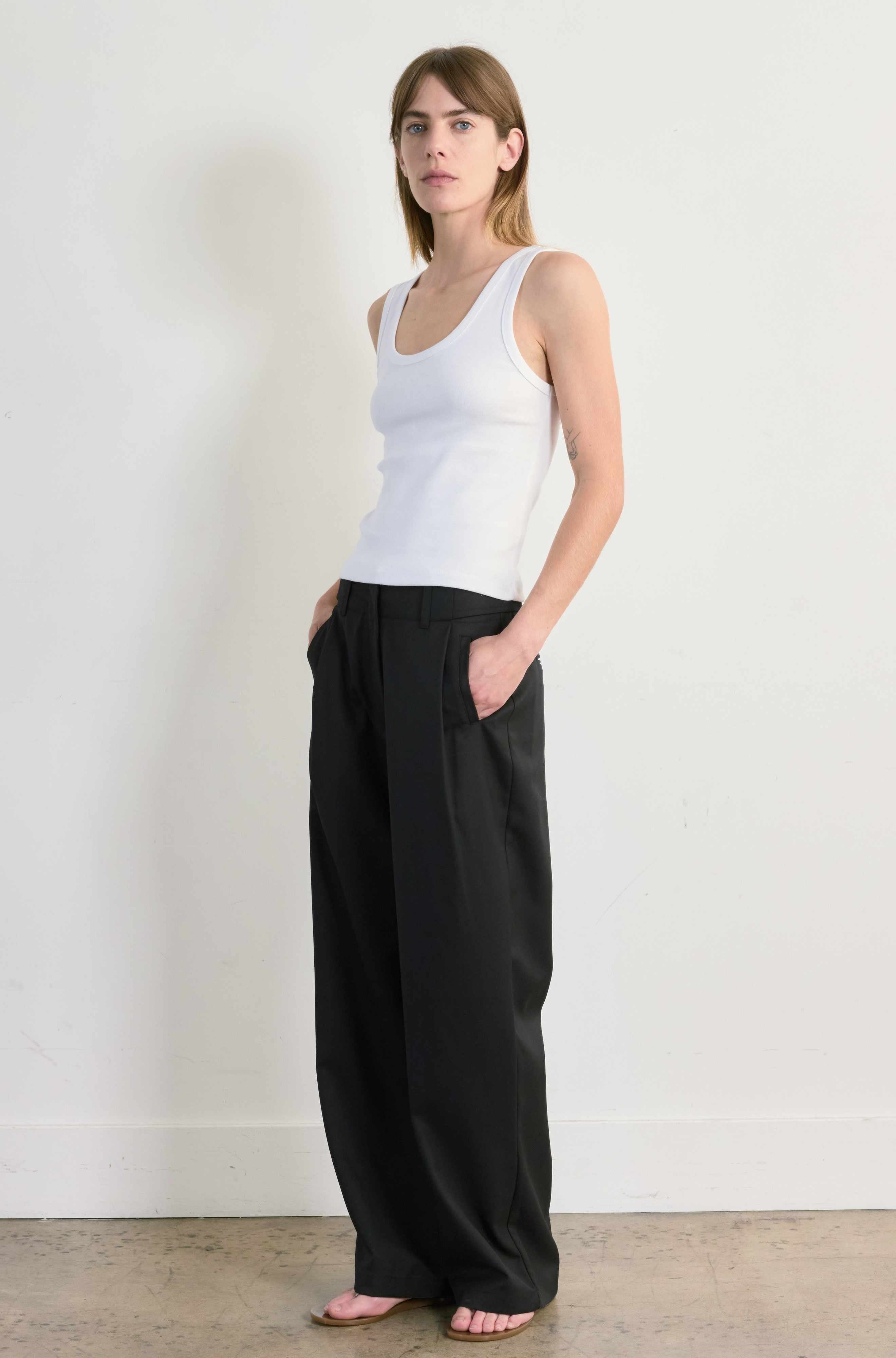 Woman wearing a white tank top and black pants against a plain background