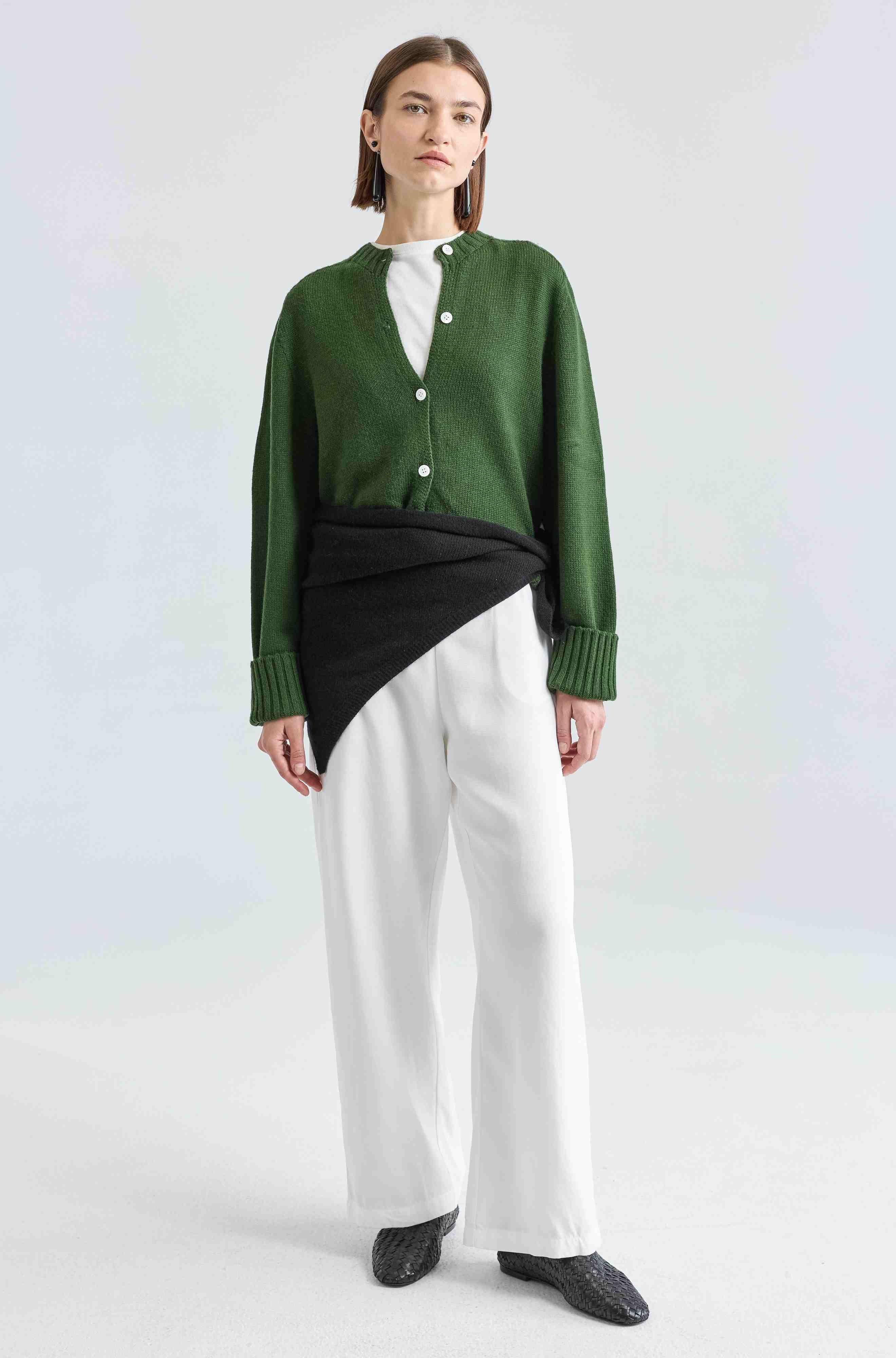 Person wearing a green cardigan over a black top with white pants on a light gray background