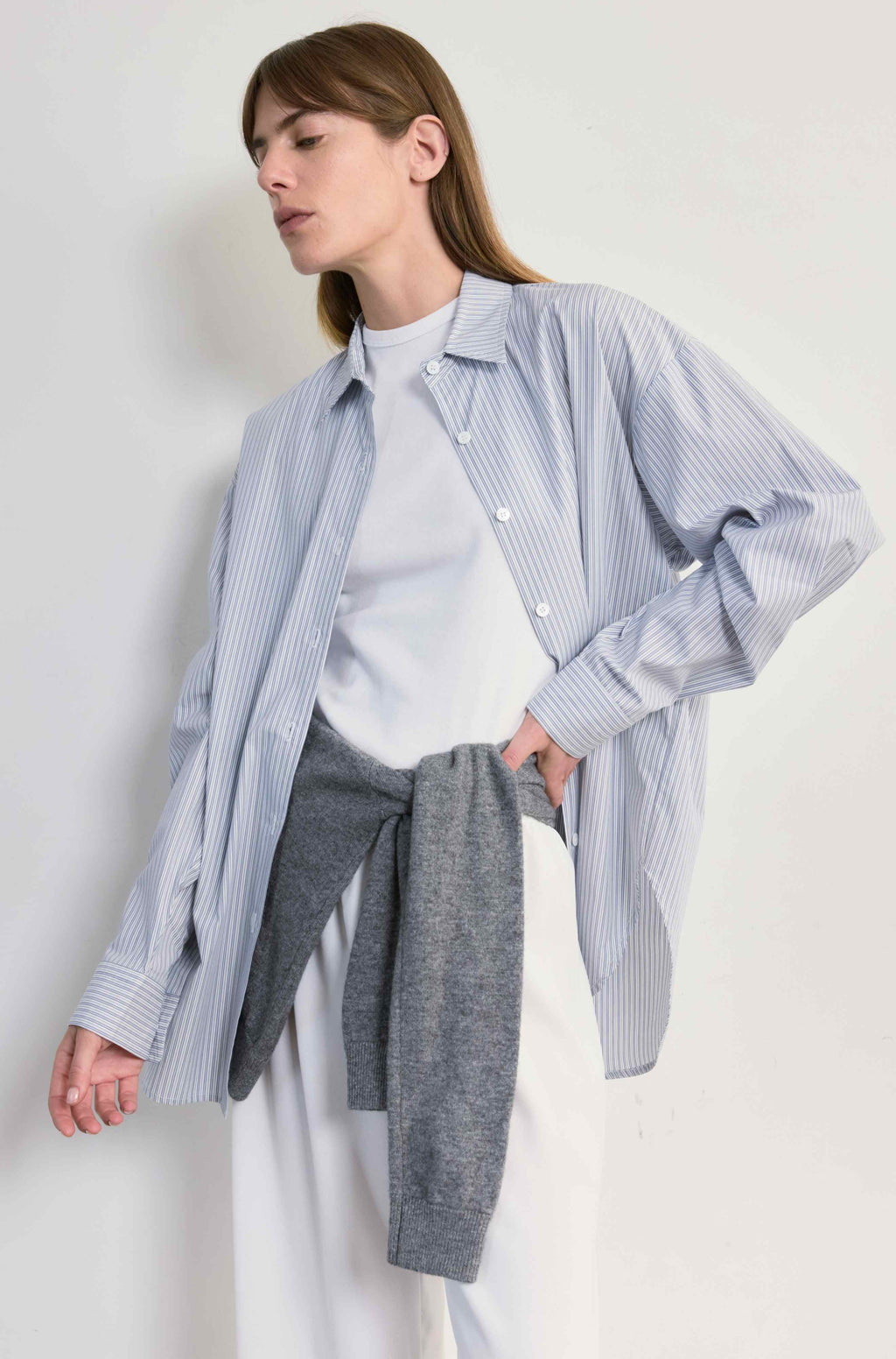 Woman wearing a light blue striped shirt over a white sweater with gray pants against a white background
