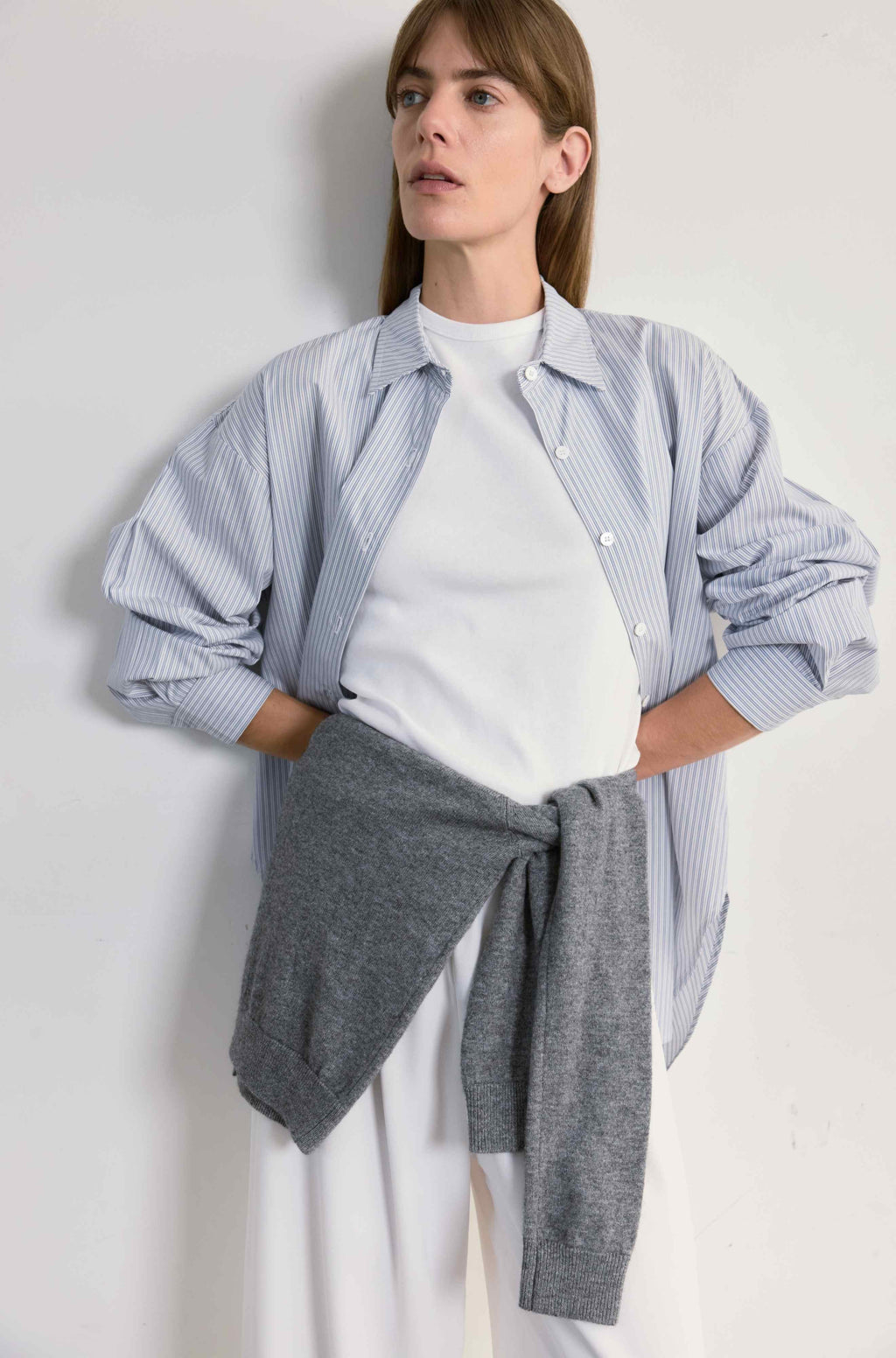 Woman wearing a light blue checkered shirt over a white top with gray pants on a plain background