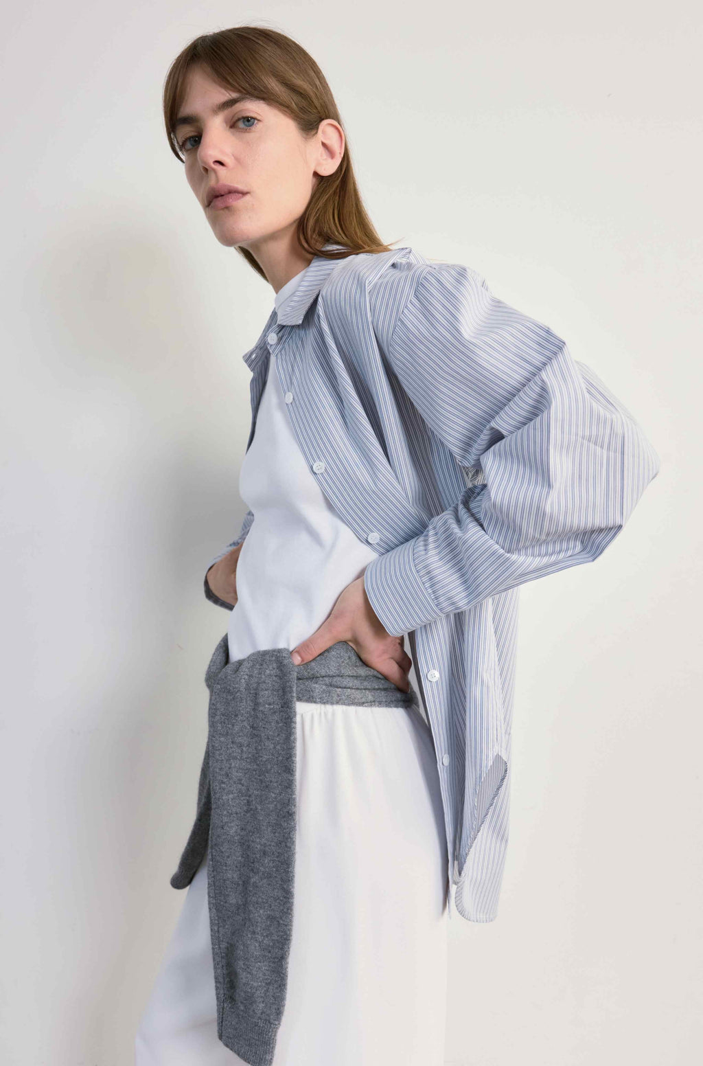 Person wearing a light blue striped shirt over a white top with a gray sweater tied around the waist, on a plain background.