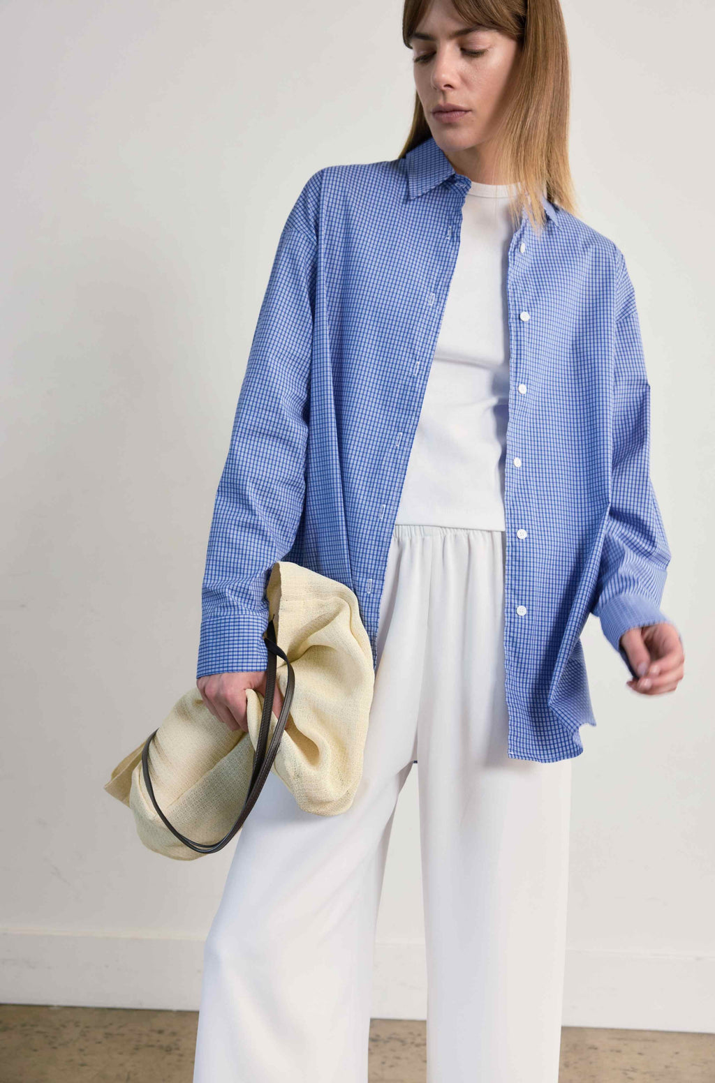 Woman wearing a blue checkered shirt over a white top with white pants, holding a beige hat.