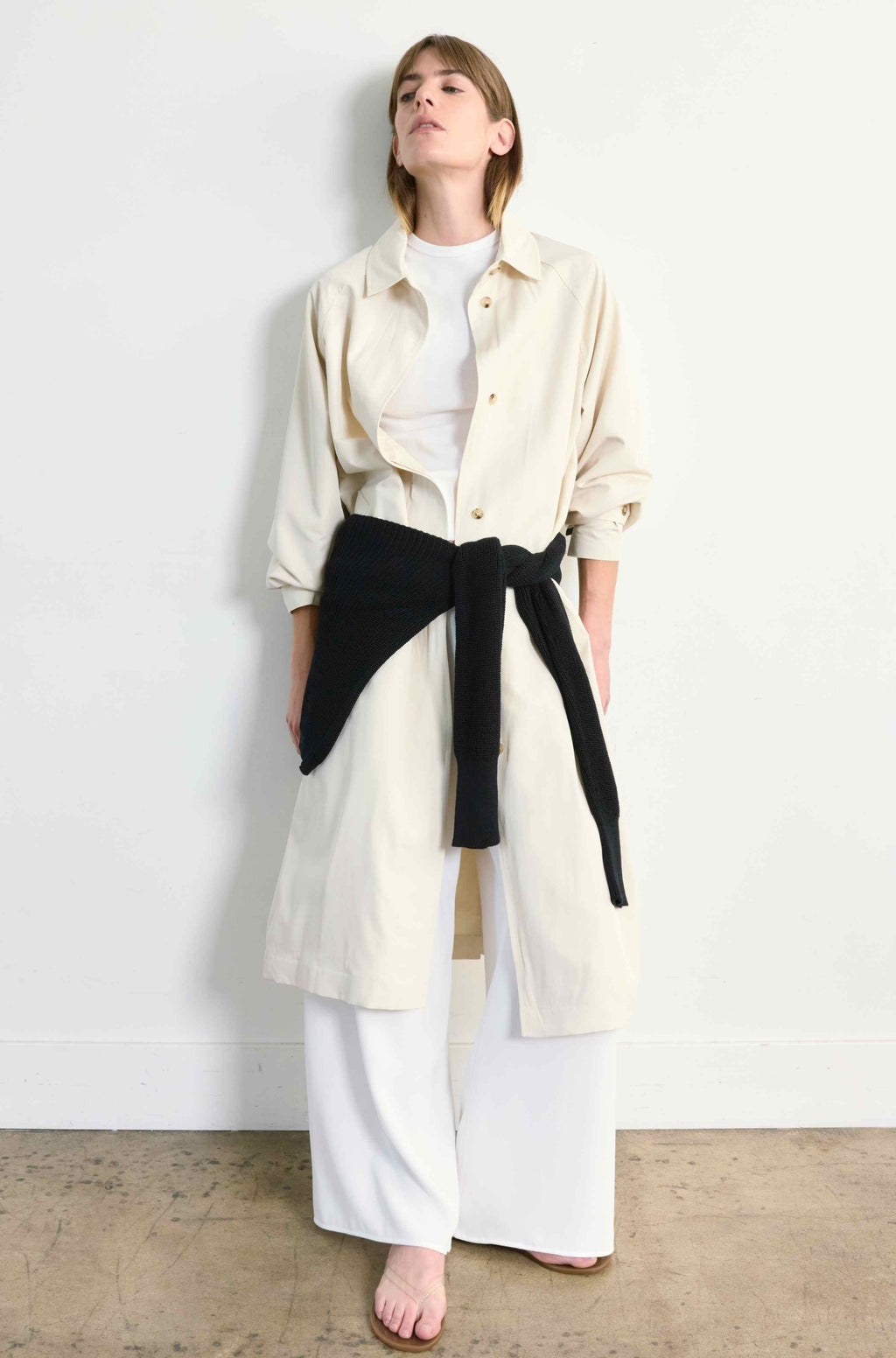 Woman wearing a long cream coat over a white outfit with a black sweater tied around her waist against a plain background
