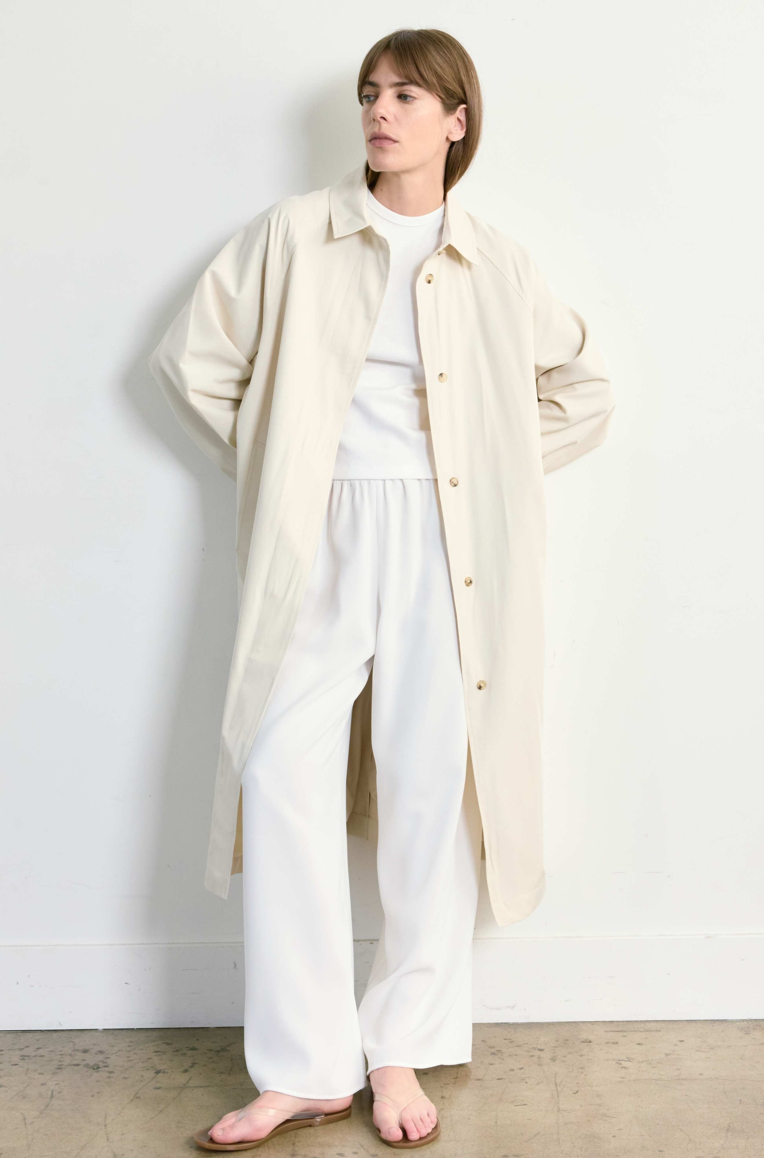 Woman wearing a long beige coat over a white outfit against a white wall.