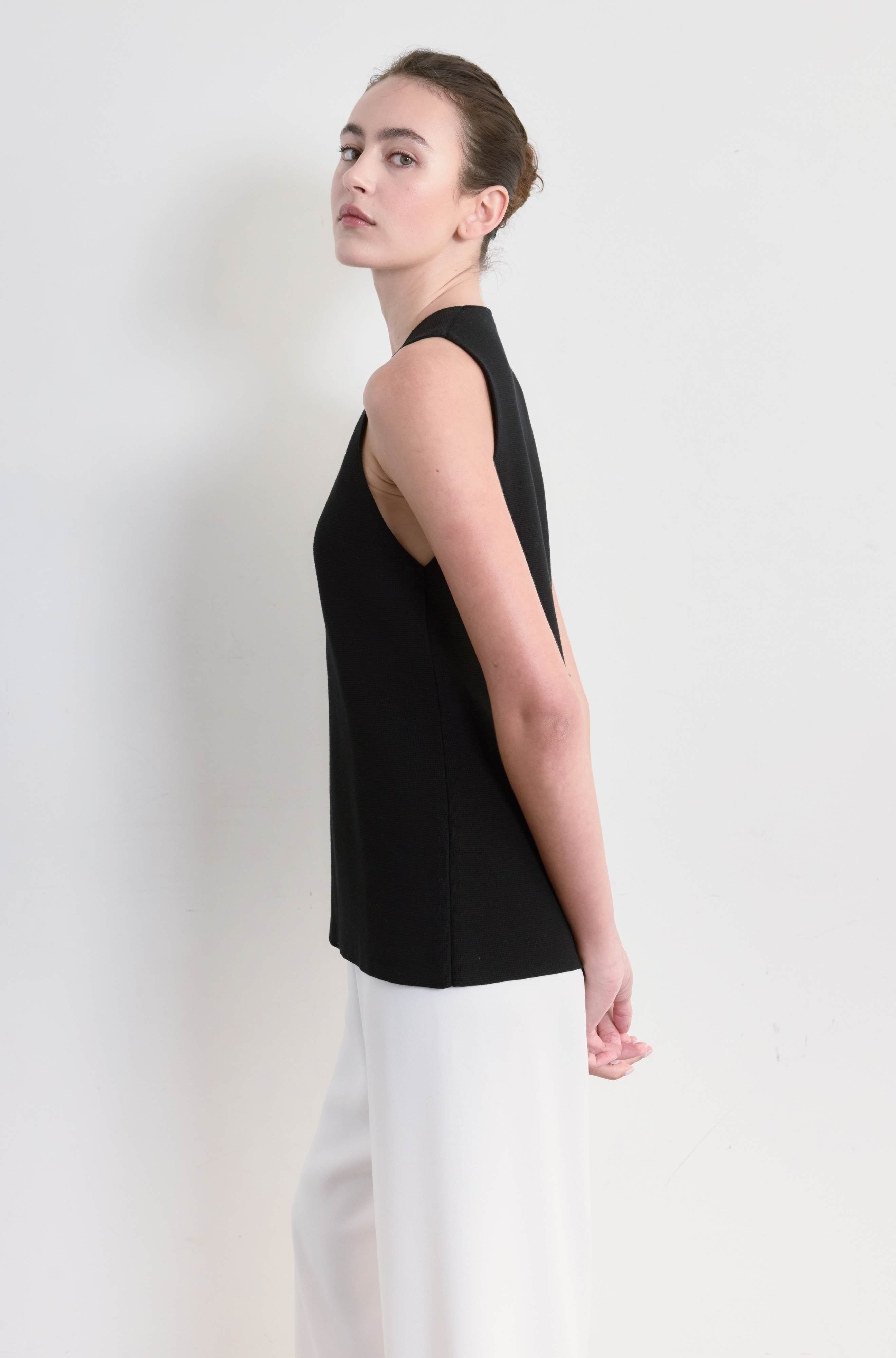 Woman wearing a black sleeveless top and white pants against a plain background