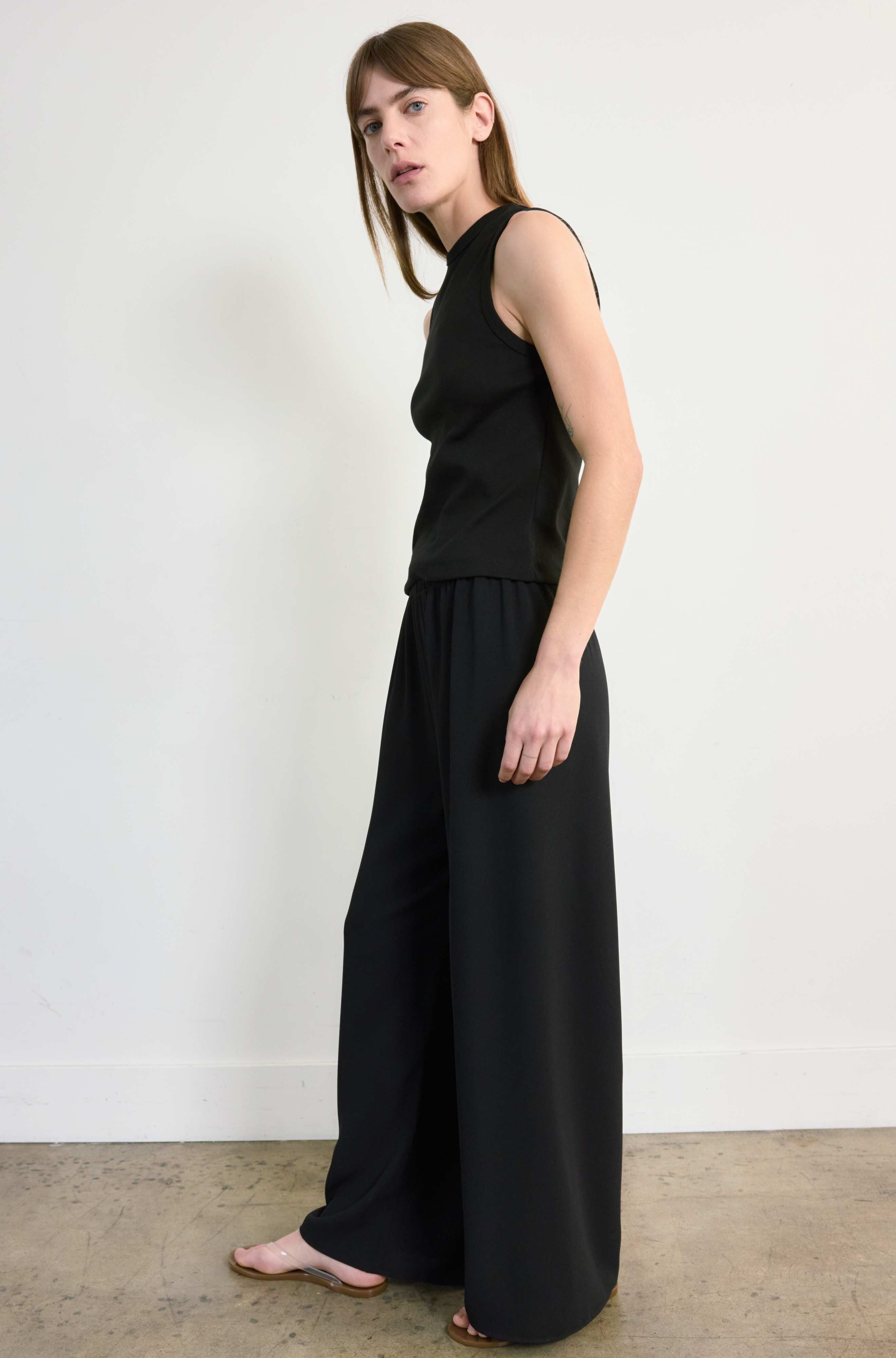 Woman wearing a black sleeveless top and wide-leg pants against a white wall.