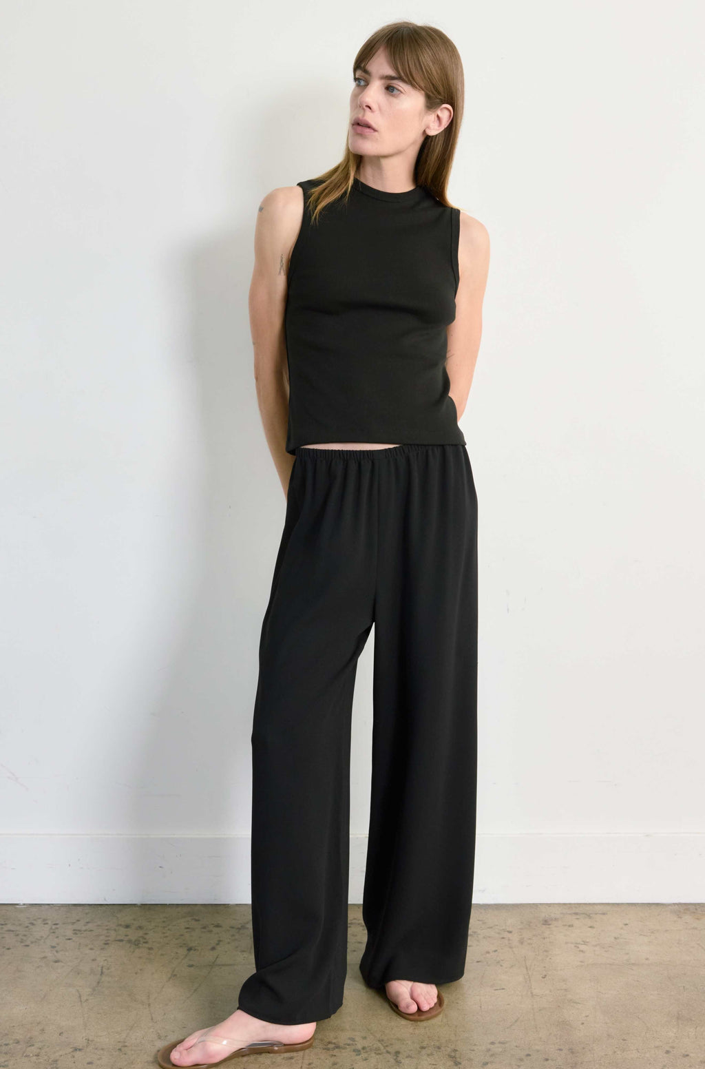 Woman wearing a black sleeveless top and wide-leg pants against a white wall.