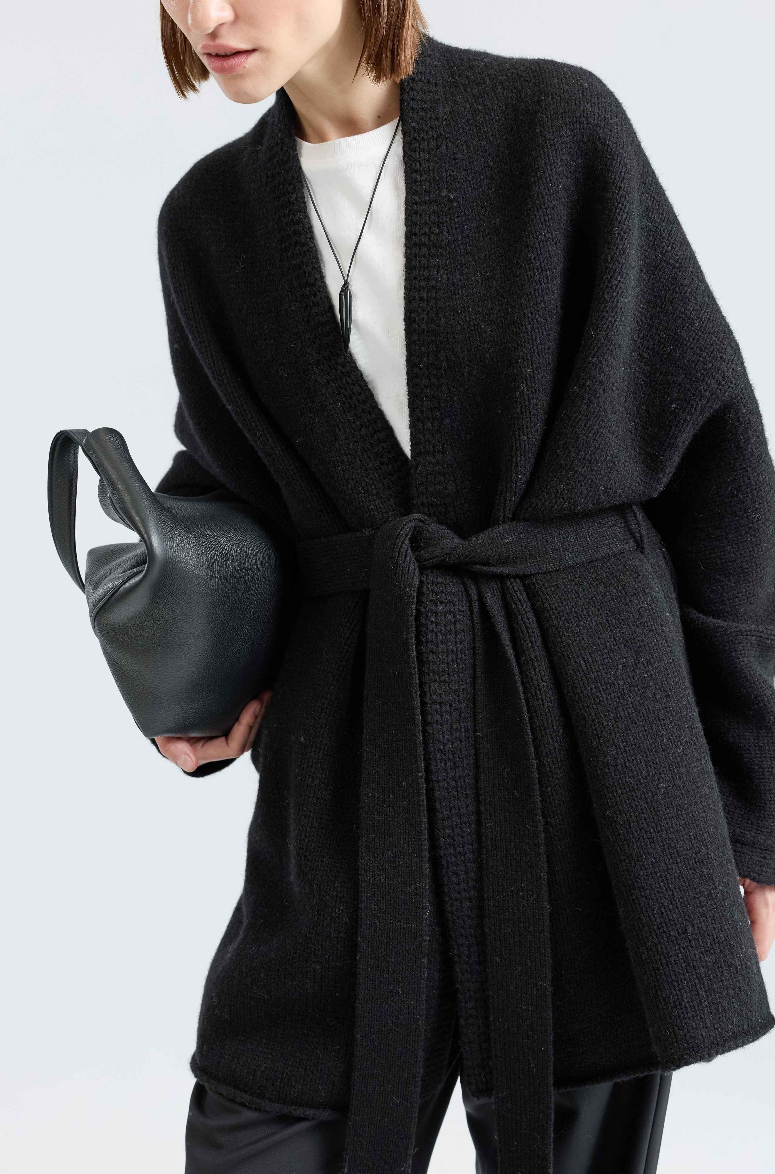 Person wearing a black coat holding a black handbag on a light gray background