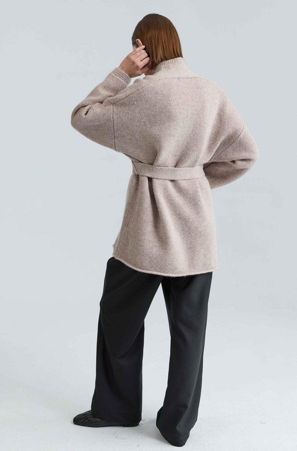 Person wearing a beige belted coat and black pants on a light gray background