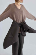 Person wearing a brown sweater and dark pants with a jacket tied around her waist against a light gray background