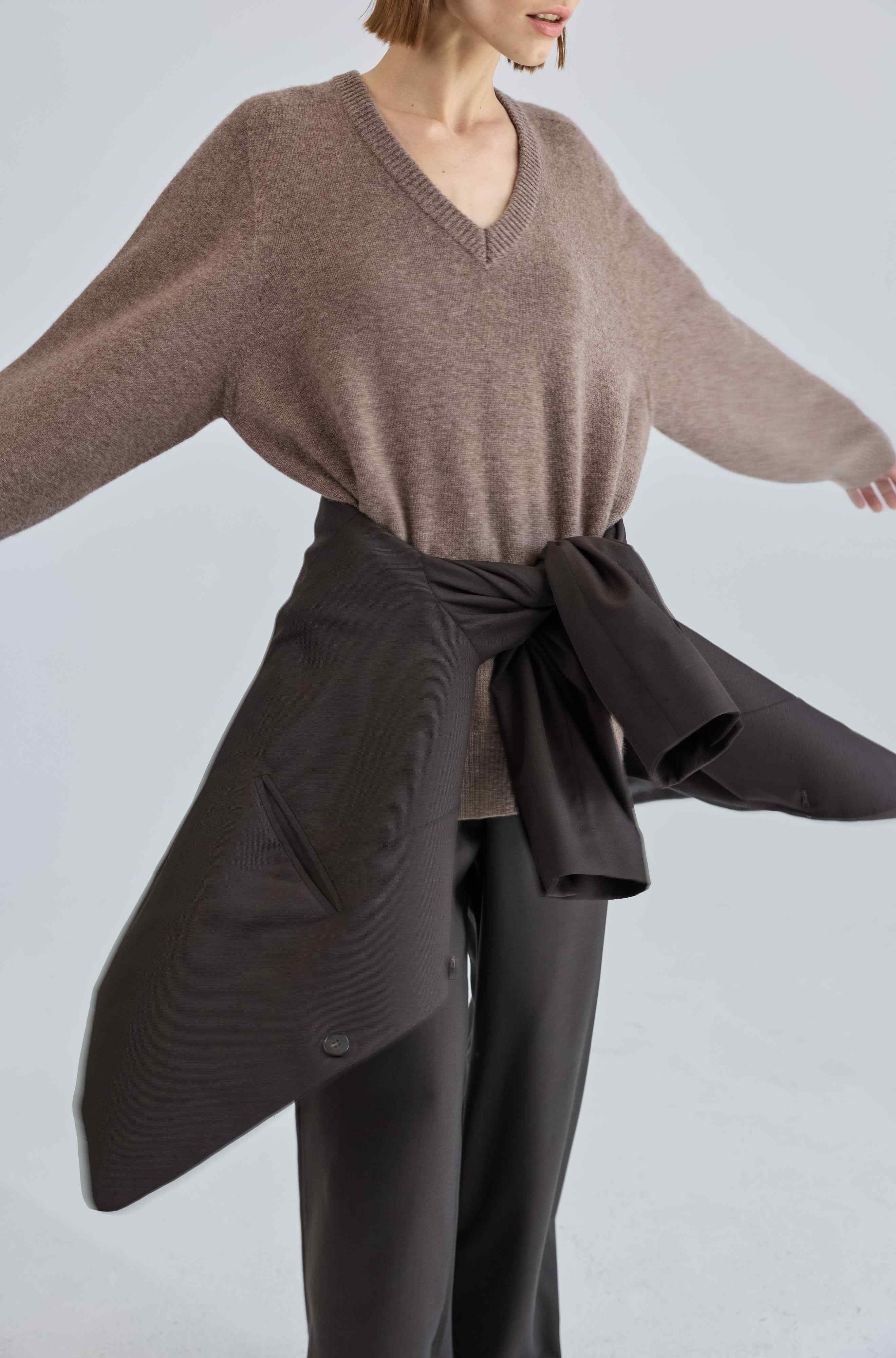 Person wearing a brown sweater and dark pants with a jacket tied around her waist against a light gray background