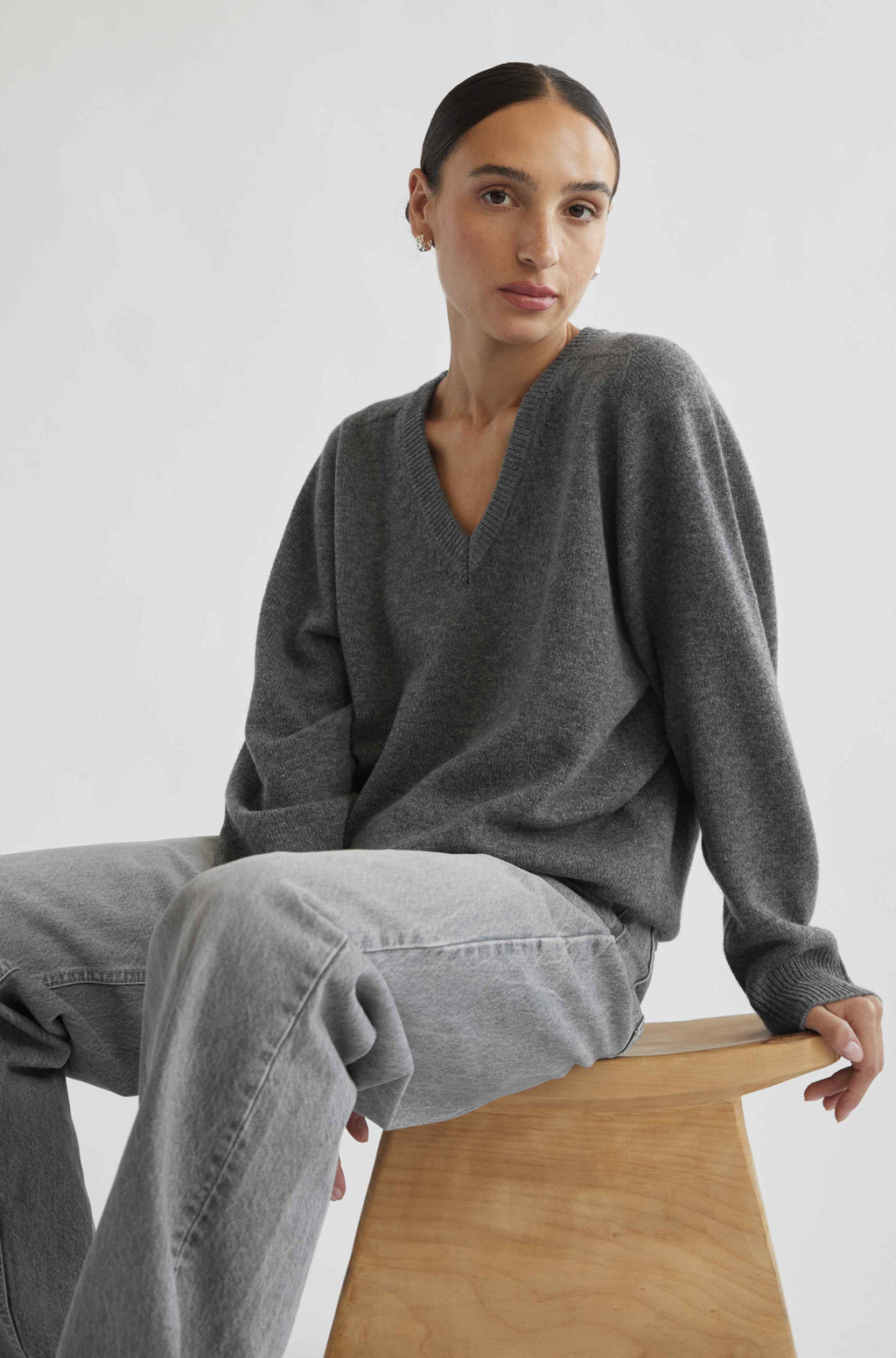 Woman wearing a gray sweater and jeans sitting on a wooden stool against a white background