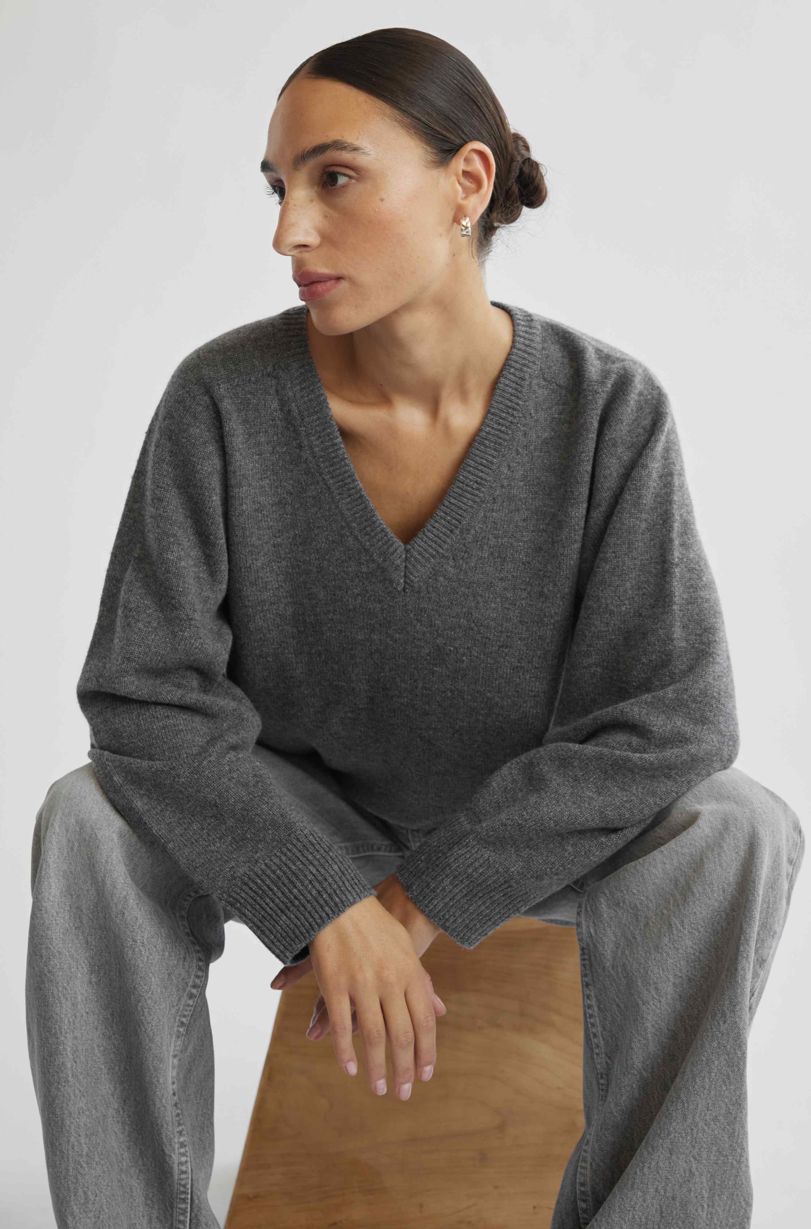 Woman wearing a gray sweater and gray pants sitting on a wooden stool against a white background