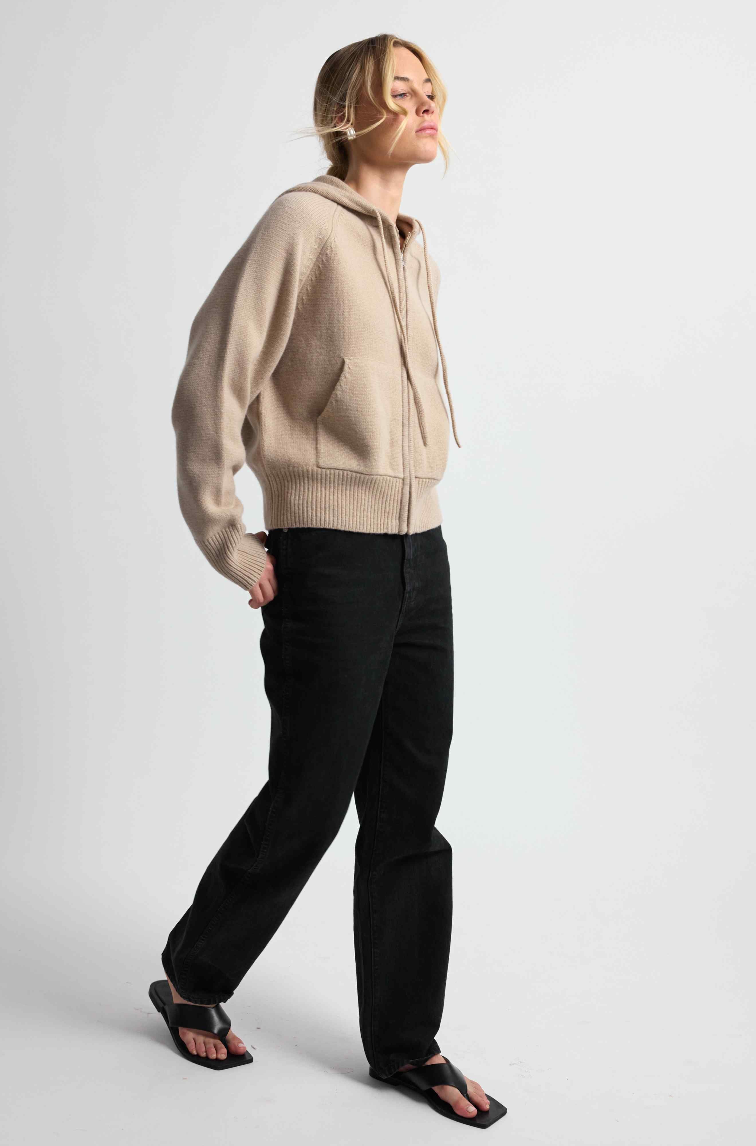 Person wearing a beige zip-up hoodie and black pants on a white background
