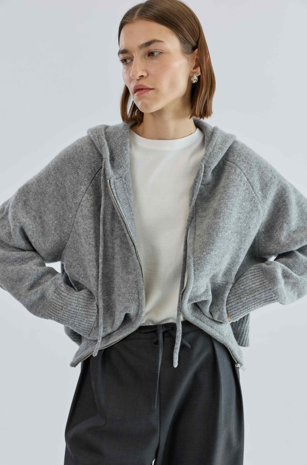 Woman wearing a gray zip-up hoodie over a white shirt and black pants on a light gray background