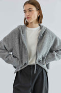 Woman wearing a gray zip-up hoodie over a white shirt and black pants on a light gray background