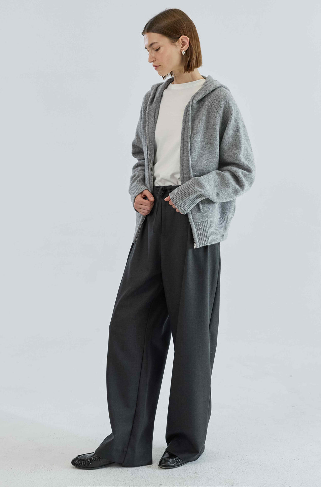 Person wearing a gray zip up sweater, white shirt, and black pants on a white background