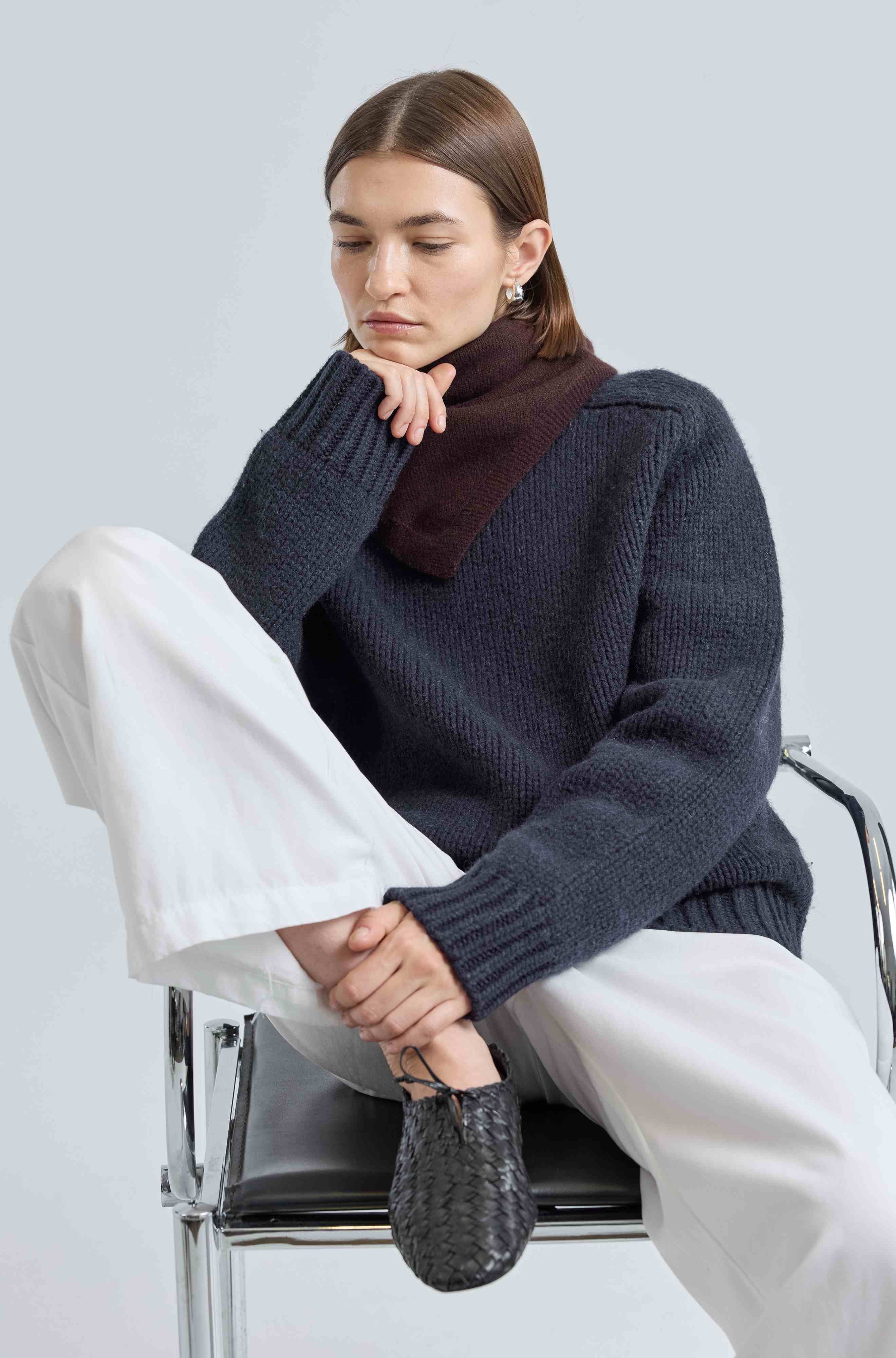Woman sitting on a chair wearing a dark blue sweater and white pants against a light gray background