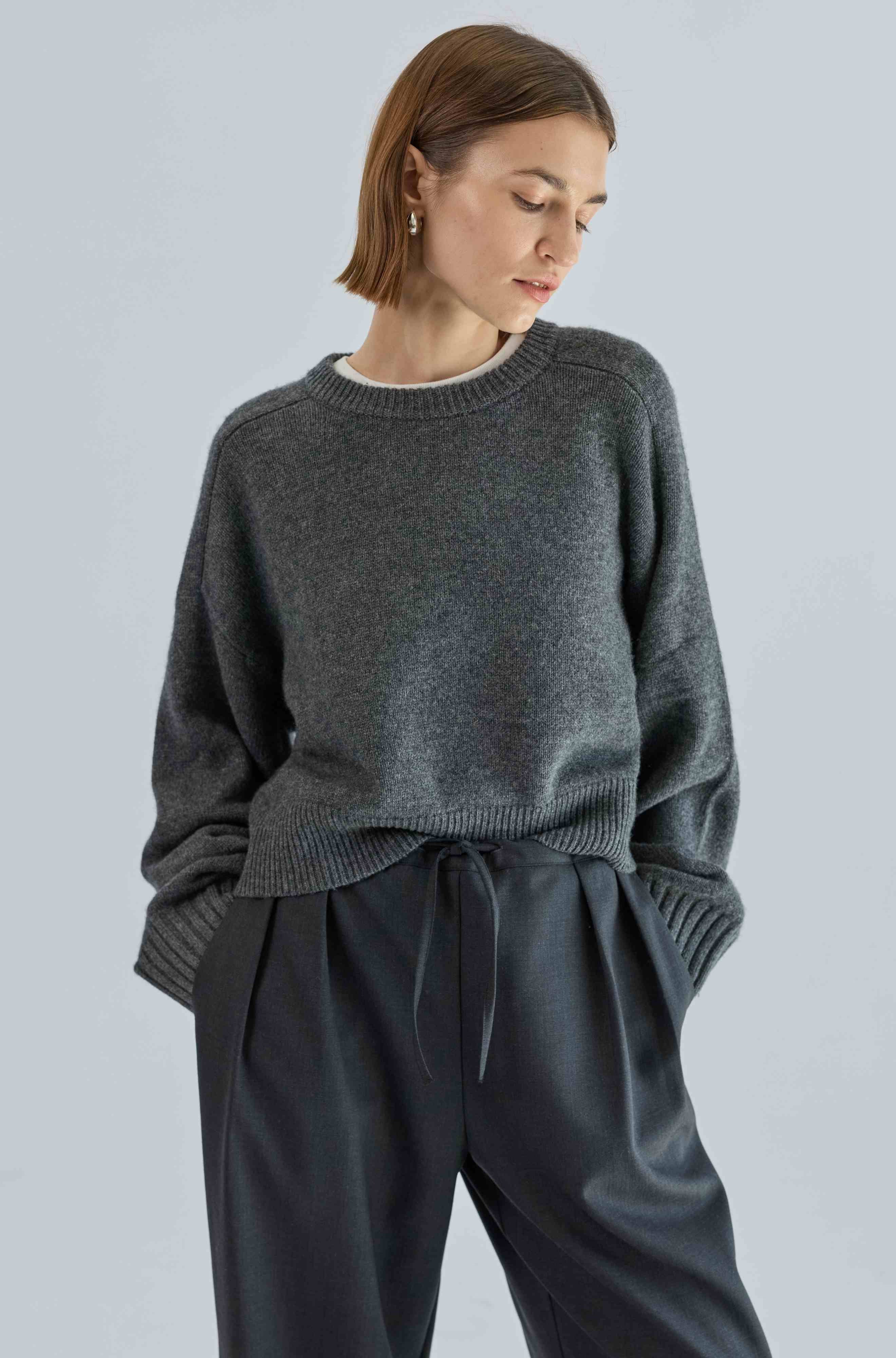 Woman wearing a gray sweater and black pants against a light gray background