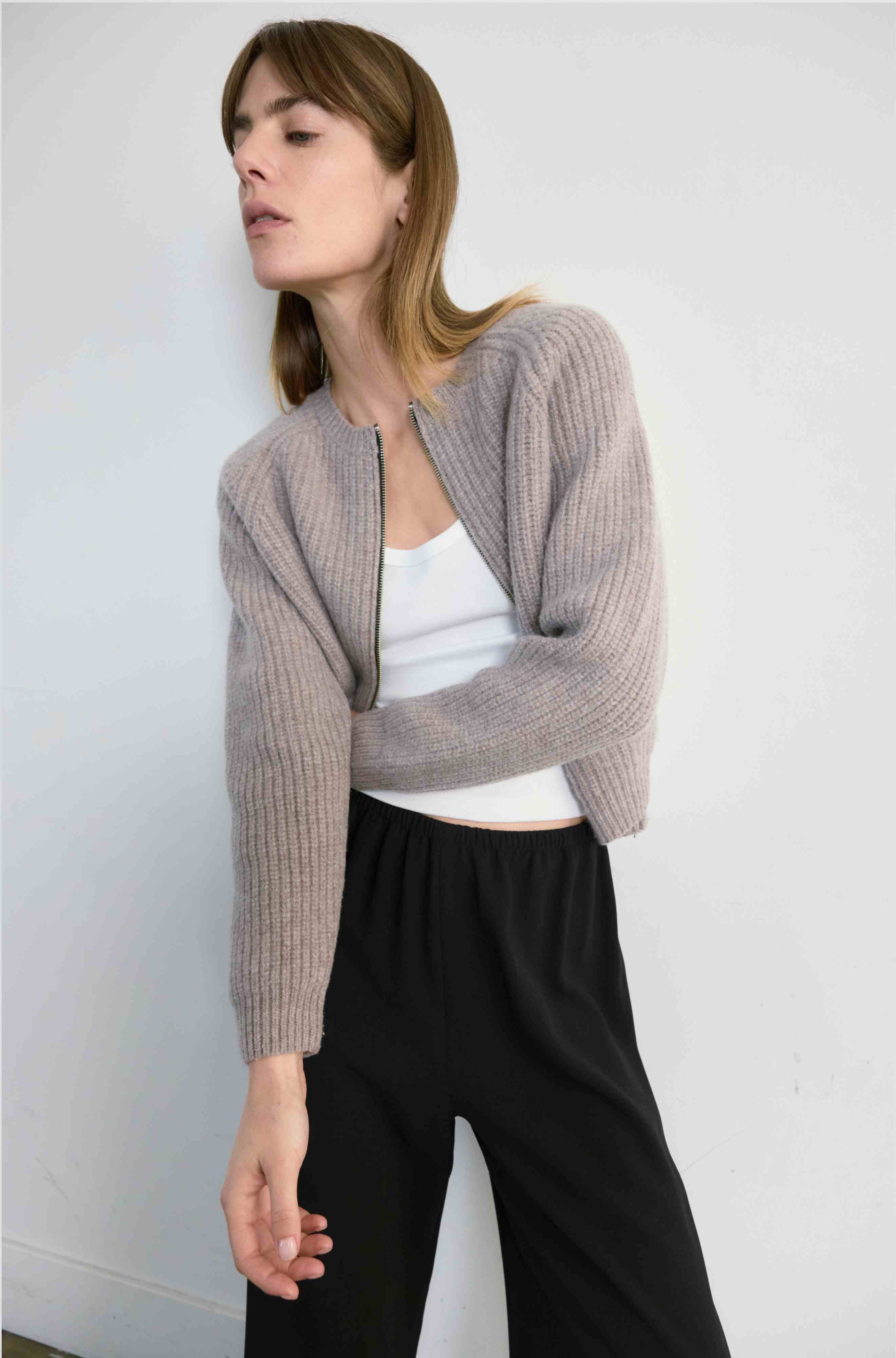 Woman wearing a light beige cardigan over a white top and black pants against a plain background