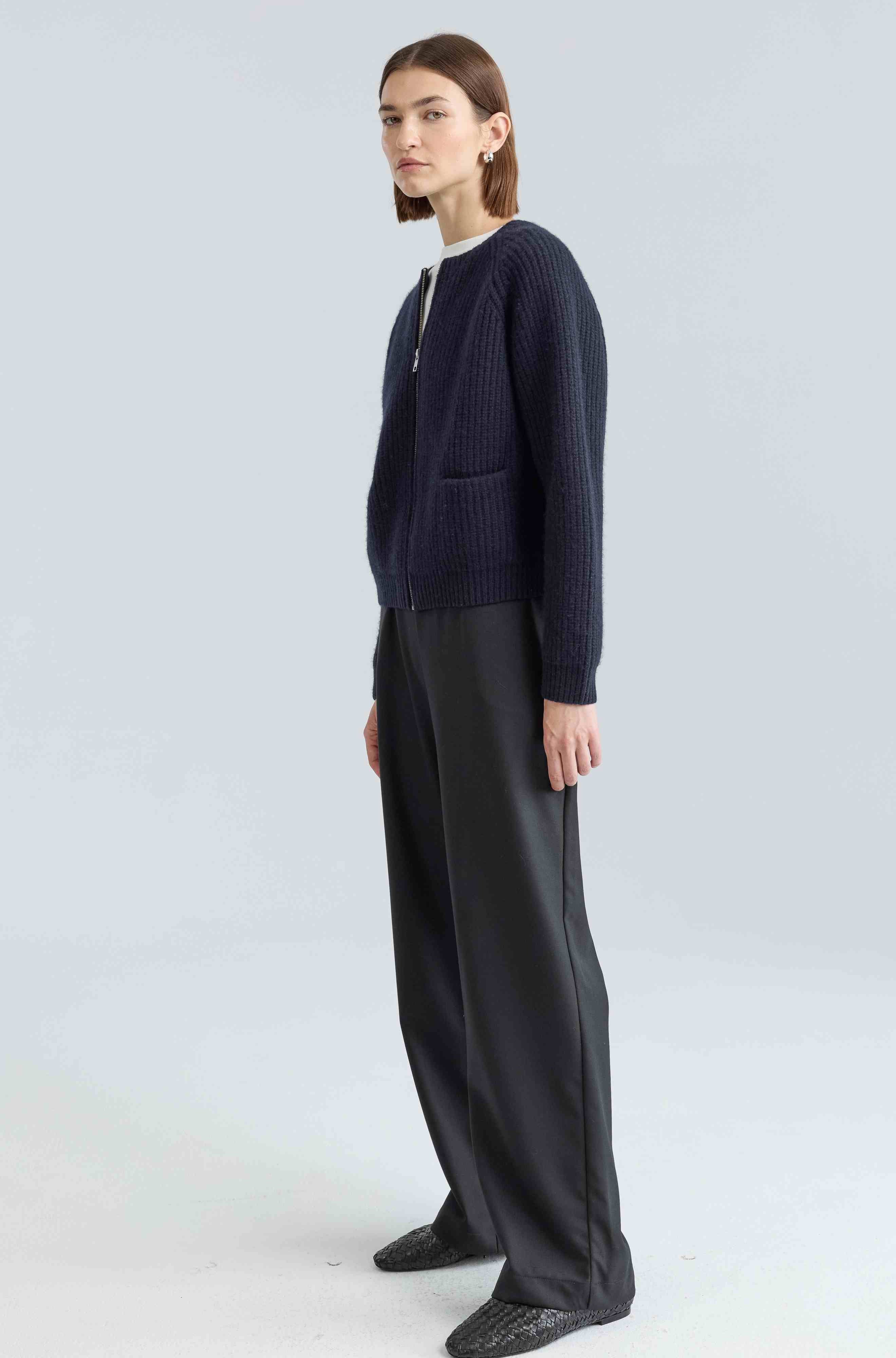 Woman wearing a navy zip up cardigan and black pants on a light gray background