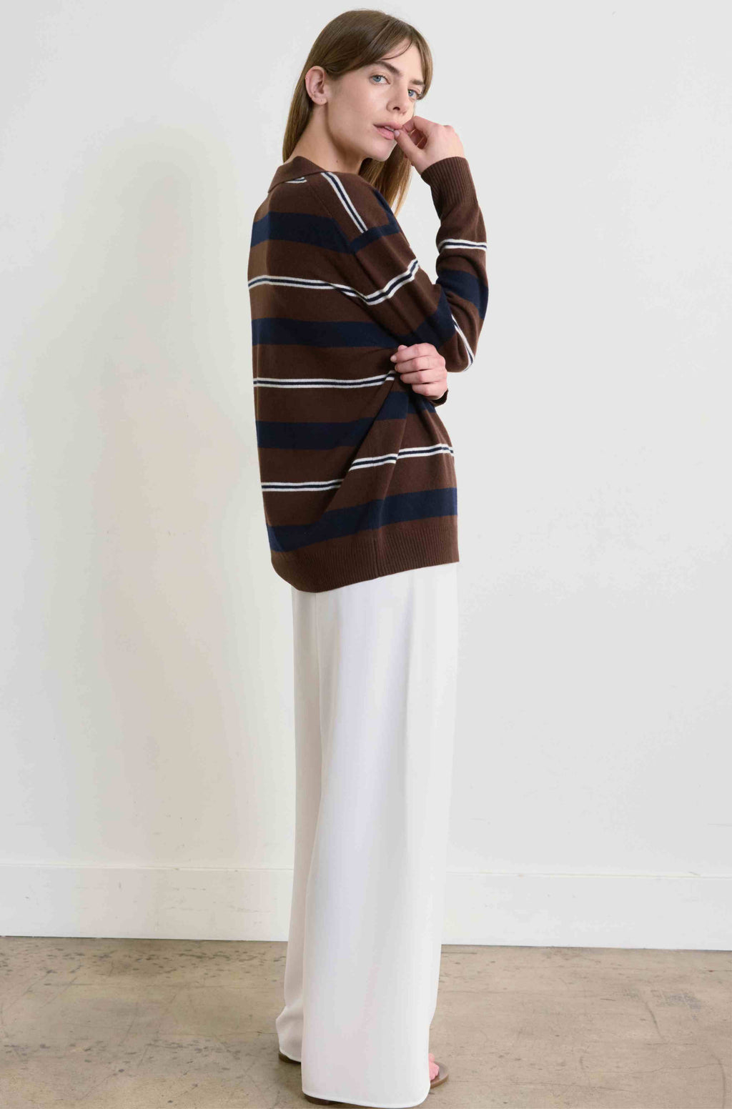 Woman wearing a brown and navy striped sweater with white pants against a plain background