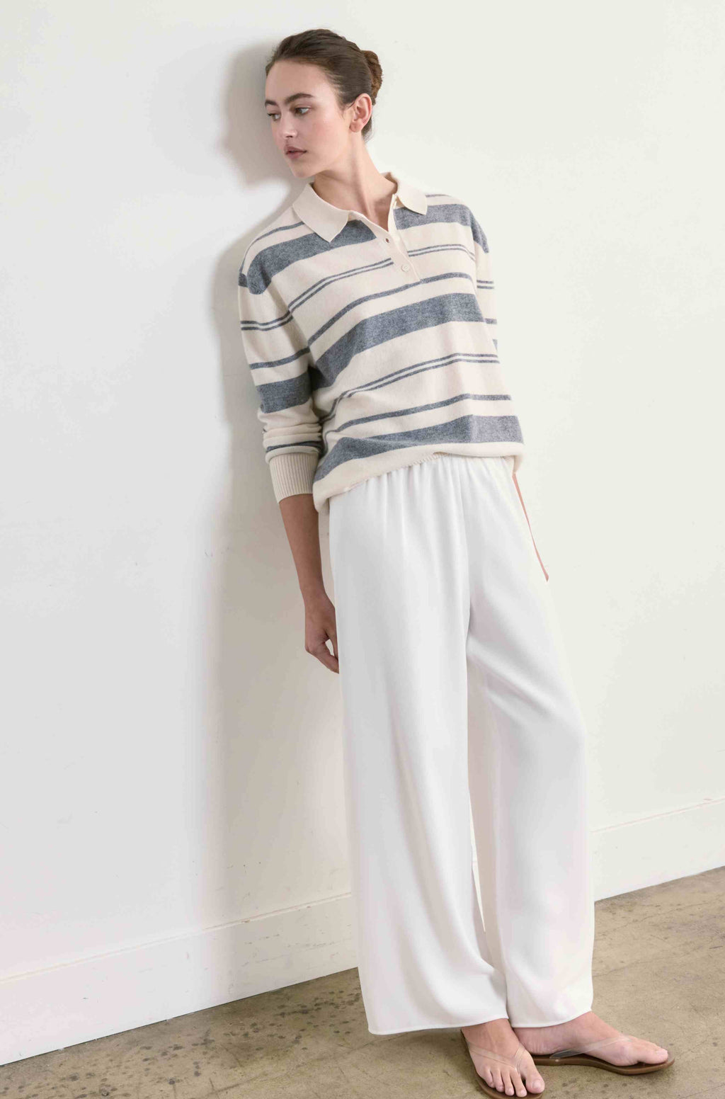 Woman wearing a striped sweater and white pants against a plain wall.