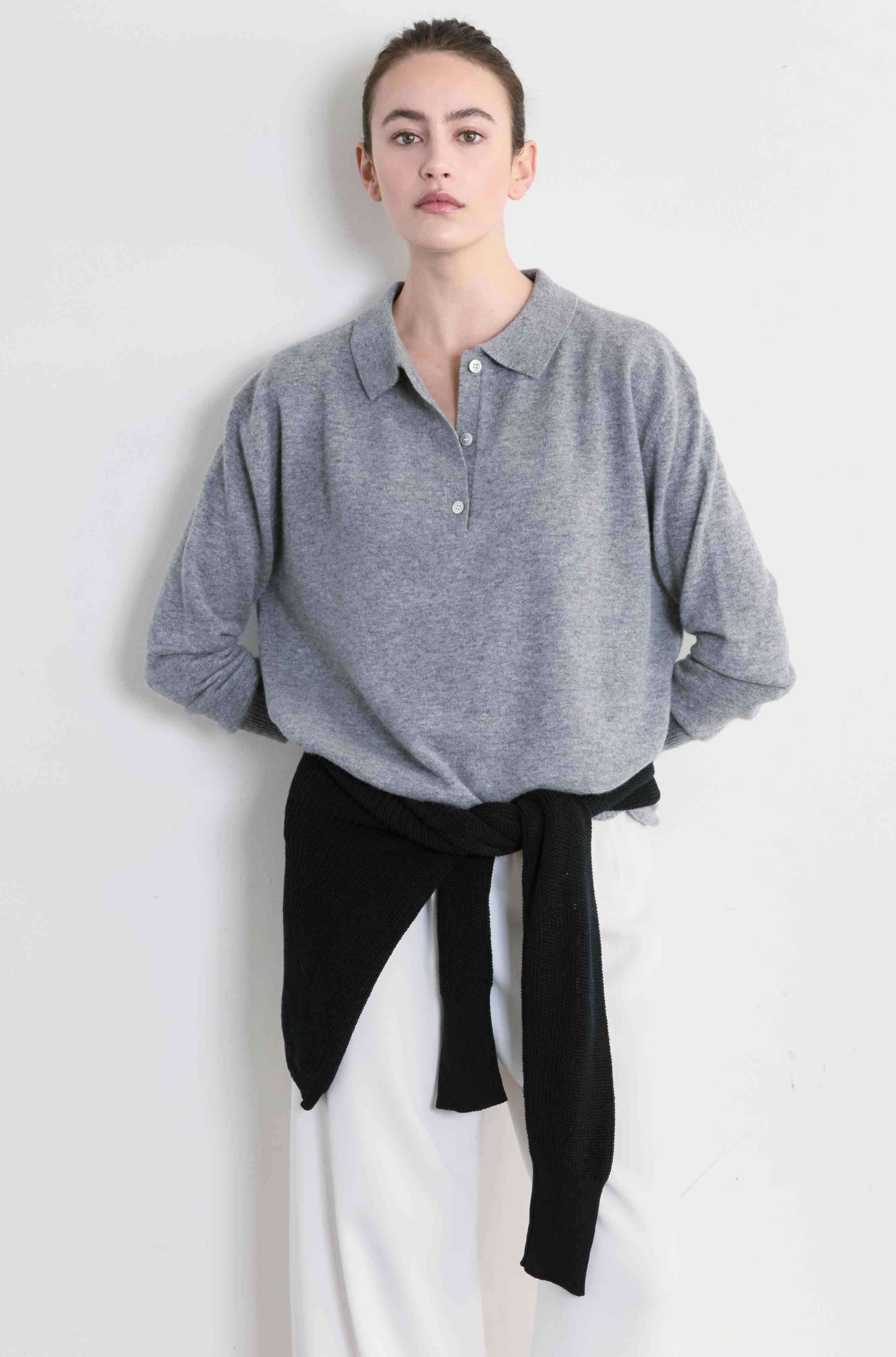Person wearing a gray sweater with a black sweater tied around her waist and white pants on a plain background