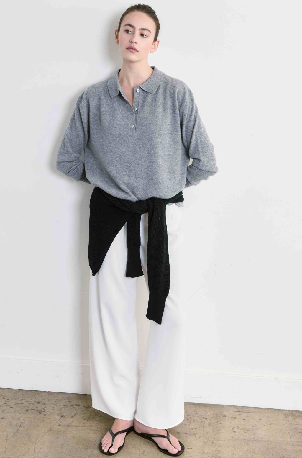 Person wearing a gray sweater, white pants, and black sweater tied around her waist against a plain background