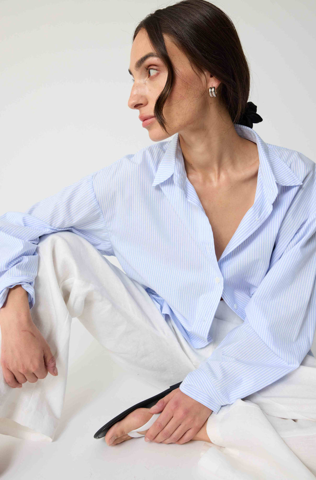 Woman wearing a light blue striped shirt and white pants sitting on a white bed.