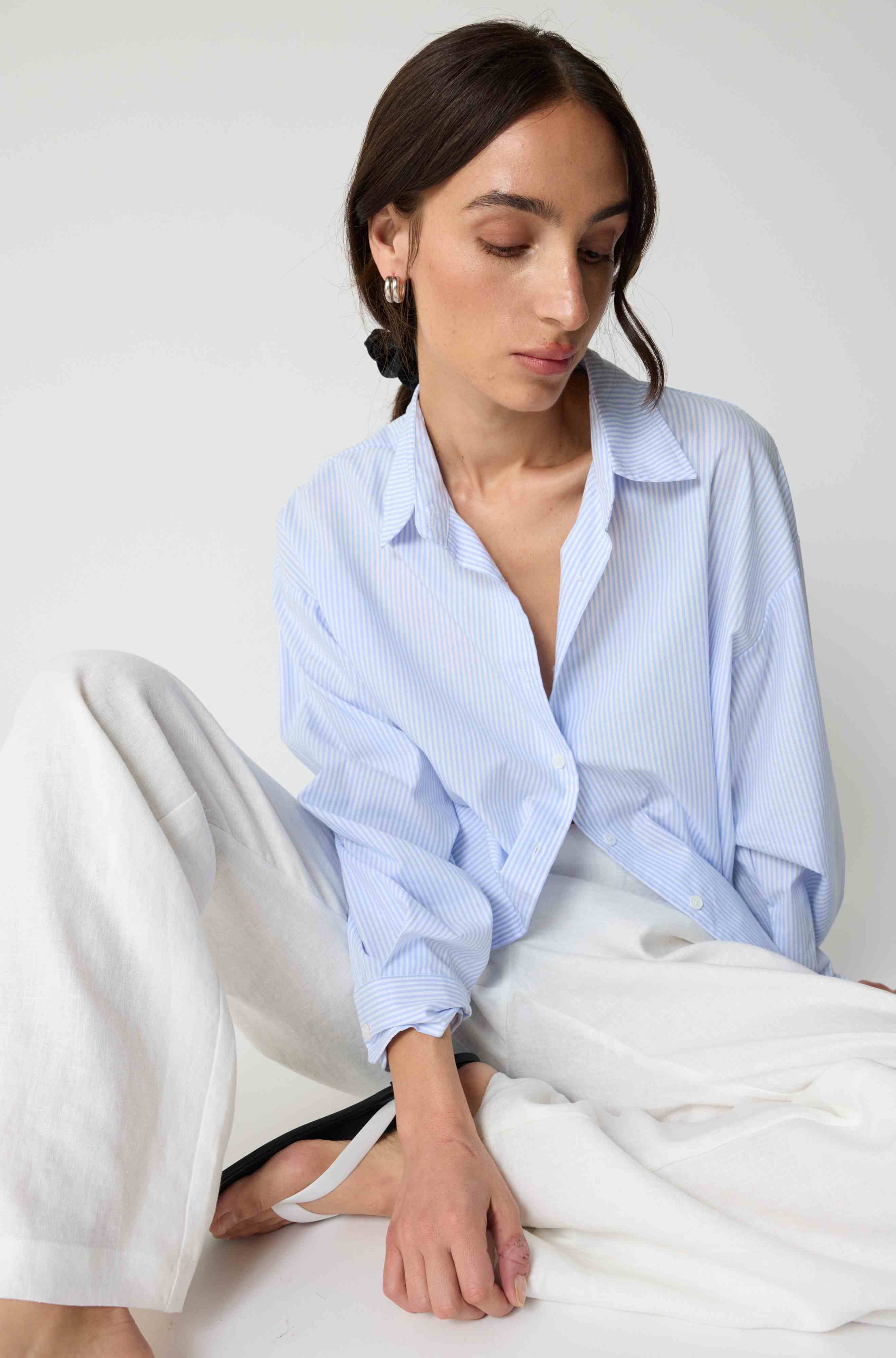Woman wearing a light blue striped shirt and white pants sitting on a bed.