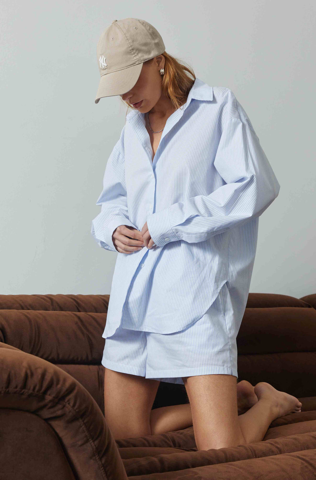 Person wearing a light blue oversized shirt and beige cap, standing on a brown couch.
