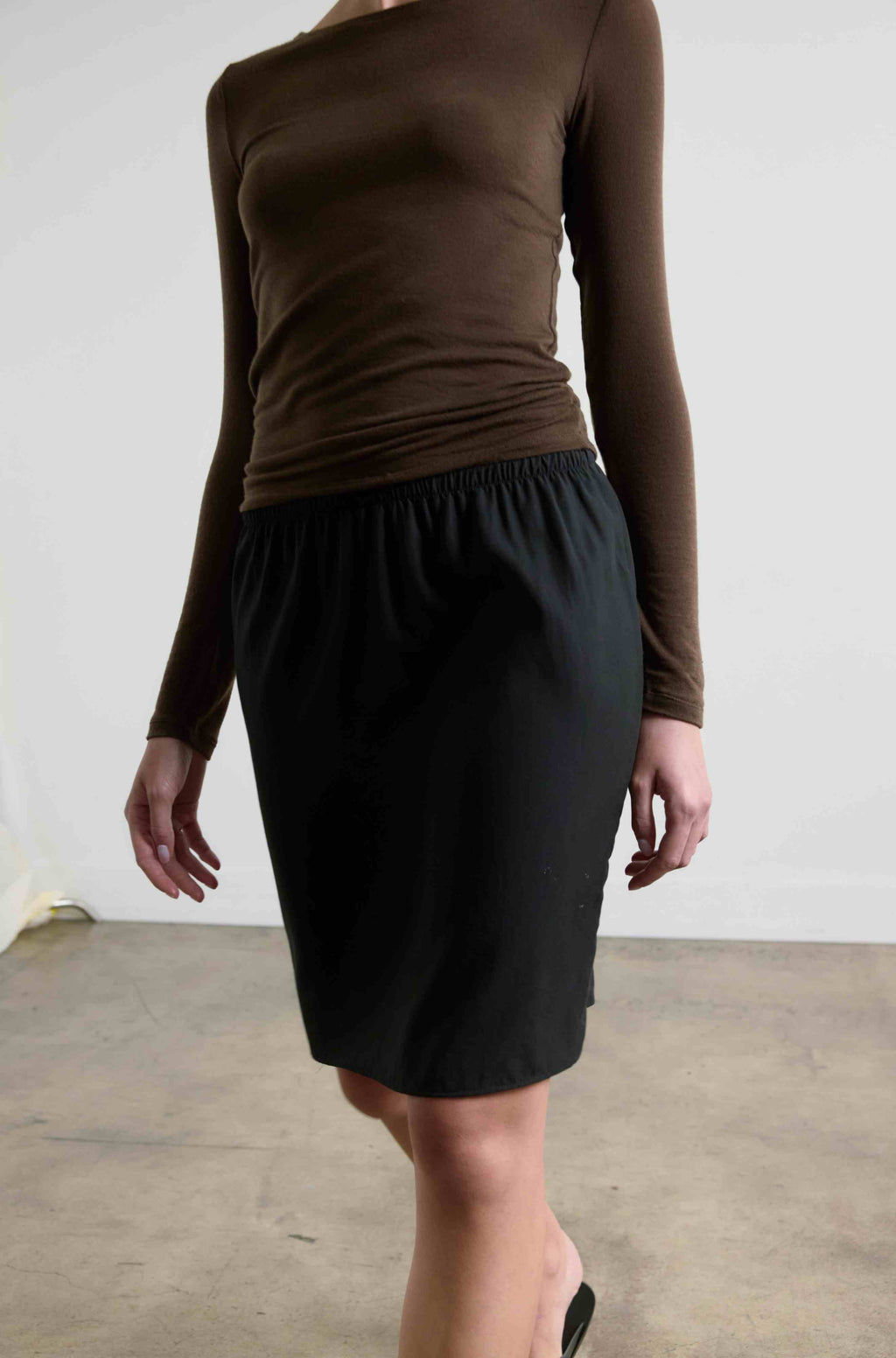 Person wearing a brown long-sleeve top and black skirt against a white wall.